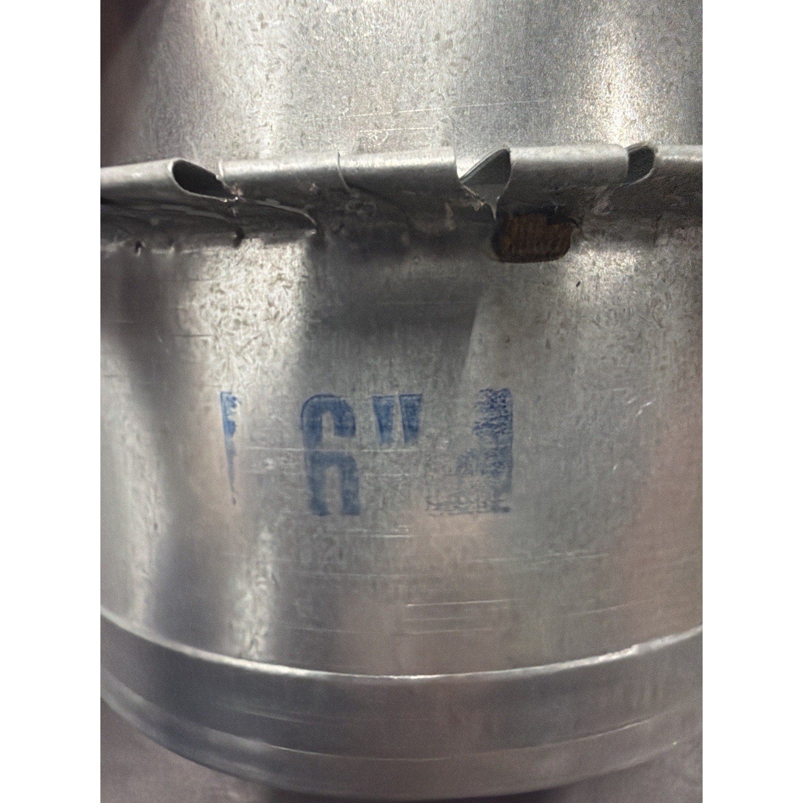 Close-up of a galvanized metal duct section showing stamped blue text indicating a 6-inch diameter with a seam and locking tabs on a reflective metal surface.