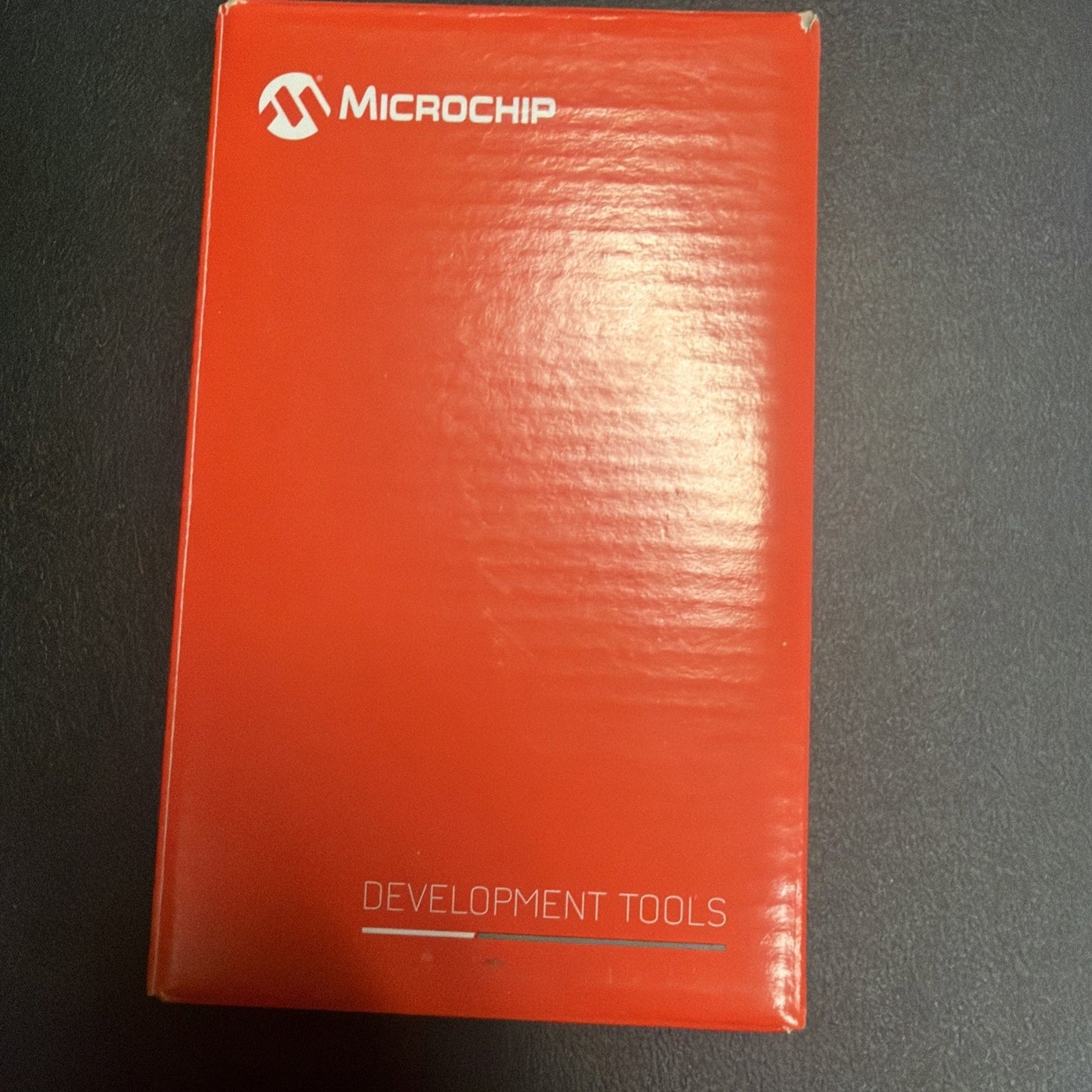 Red rectangular cardboard box with the Microchip logo and the words "Development Tools" printed on the front, placed on a dark textured surface.