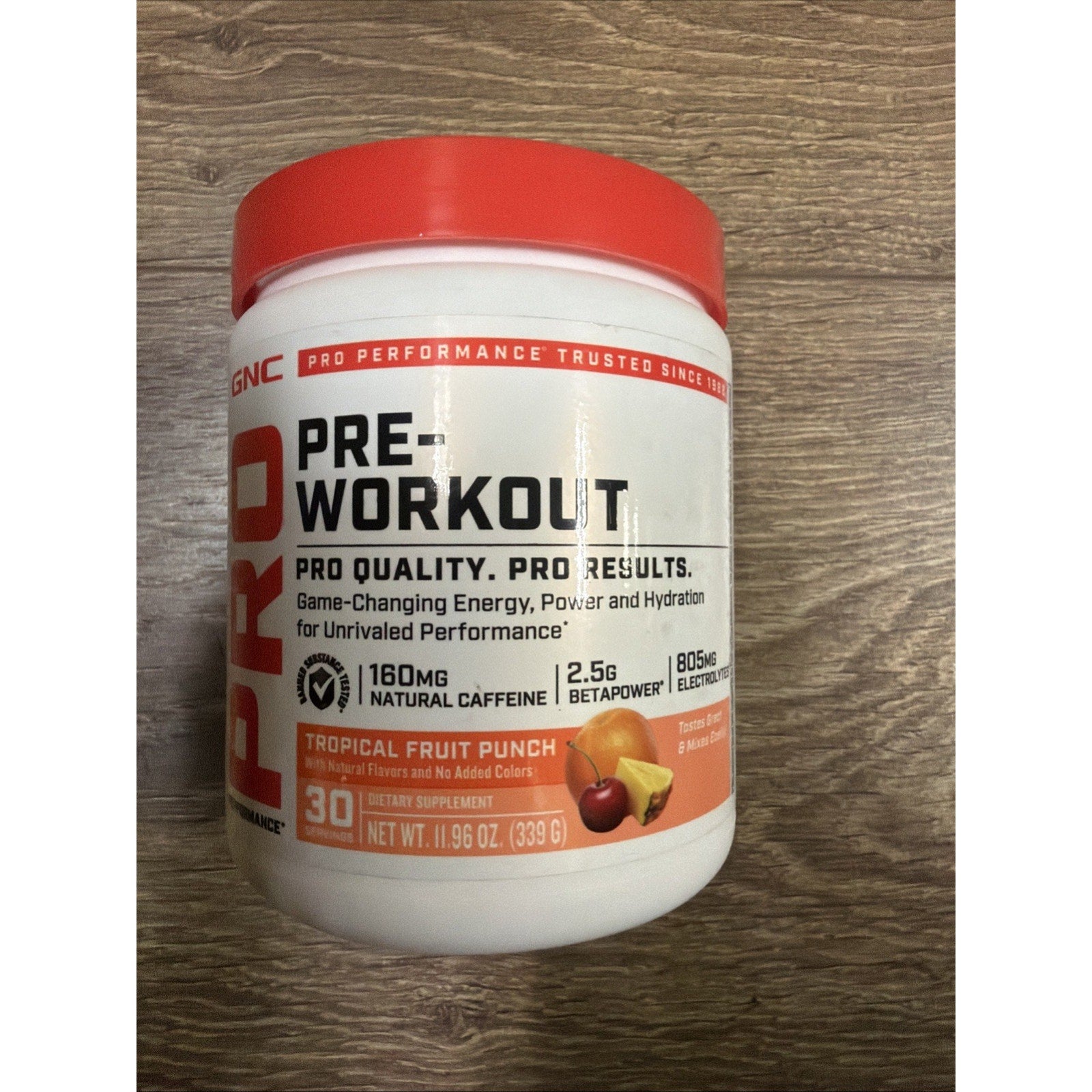 White and red container of GNC Pro Performance Pre-Workout powder in tropical fruit punch flavor, featuring natural caffeine, betapower, electrolytes, and supplement facts on a wood surface.