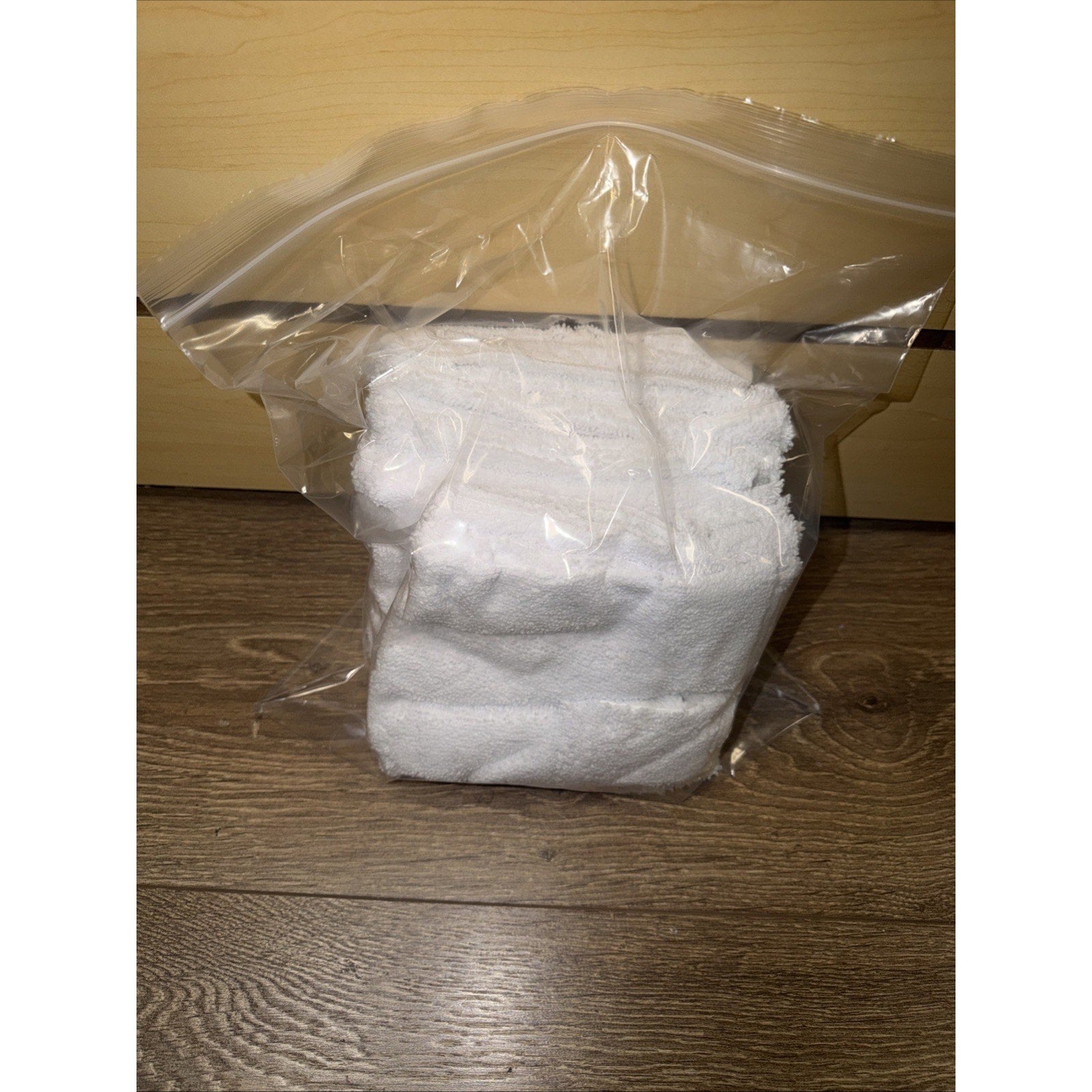 Two folded white terry cloth towels packaged inside a clear resealable plastic bag on a wooden floor with a light wood background.
