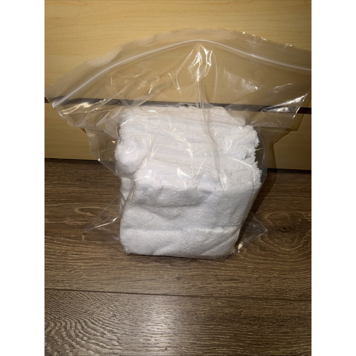 Two folded white terry cloth towels packaged inside a clear resealable plastic bag on a wooden floor with a light wood background.
