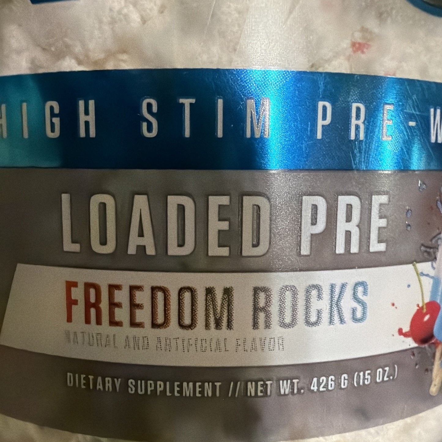 Close-up of a supplement container label showing the text "Loaded Pre," flavor name "Freedom Rocks" in red and gray, and net weight of 426 grams (15 ounces) on a textured background.