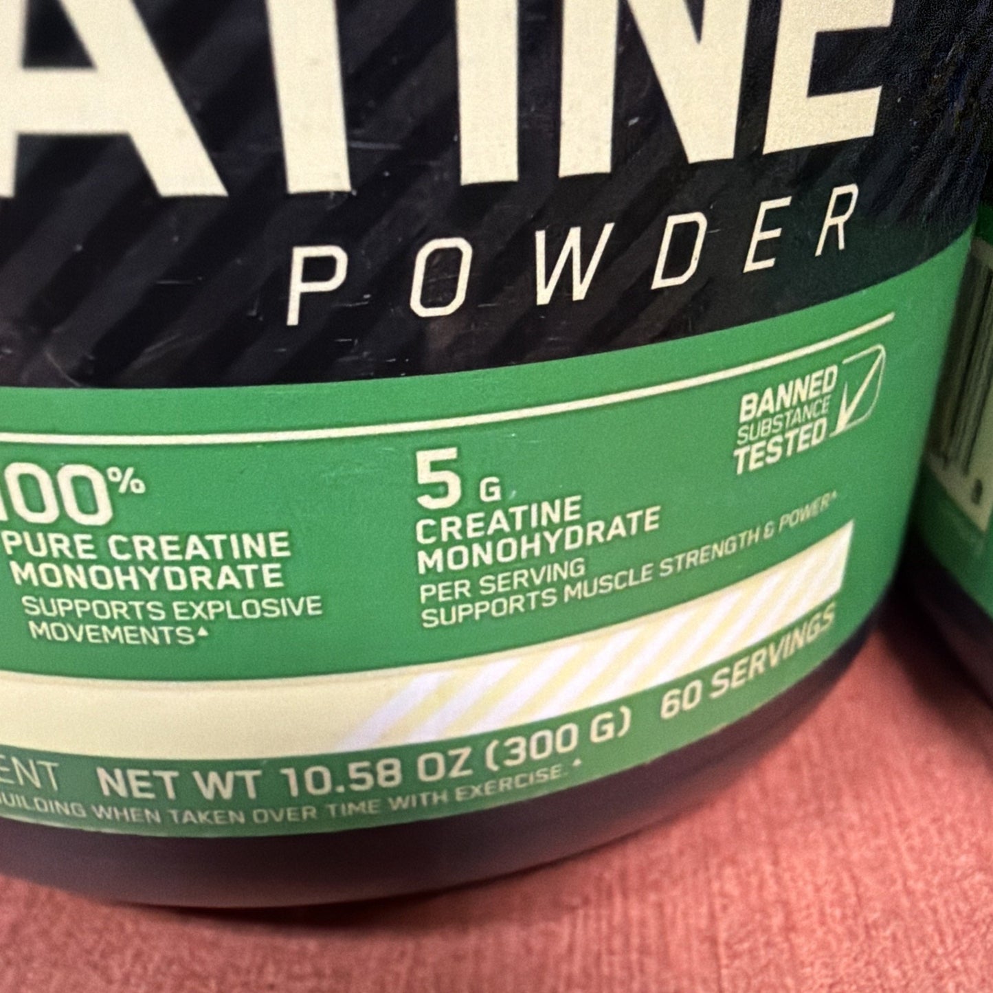 Close-up of a green label on a creatine monohydrate powder container highlighting 100% pure creatine, 5 grams per serving, 60 servings, and banned substance tested certification.