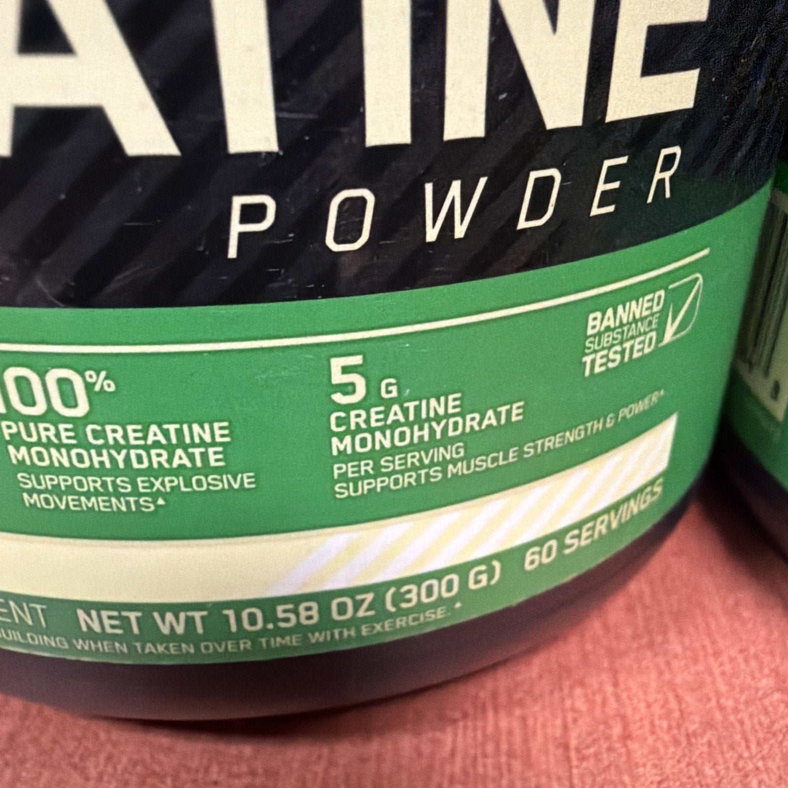 Close-up of a green label on a creatine monohydrate powder container highlighting 100% pure creatine, 5 grams per serving, 60 servings, and banned substance tested certification.