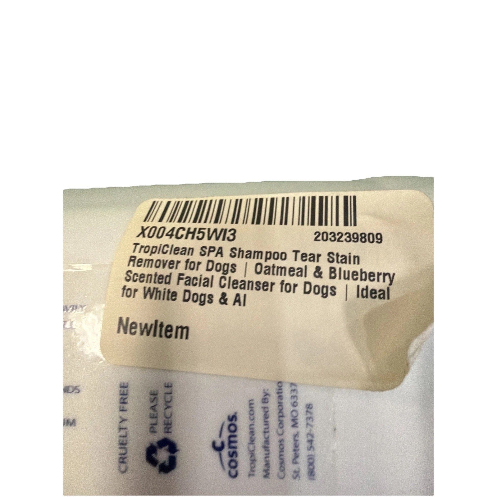 Close-up of a white product label on plastic packaging showing a barcode and printed text describing a dog shampoo tear stain remover with oatmeal and blueberry scent, designed for white dogs and AI.