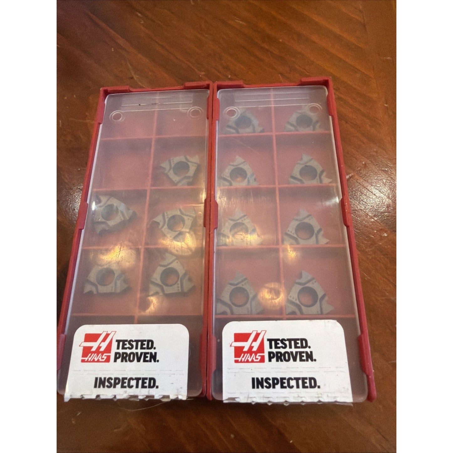 Red plastic cases with transparent lids each containing ten triangular metal cutting inserts, labeled with Haas logo and text "Tested. Proven. Inspected." on a wooden surface.