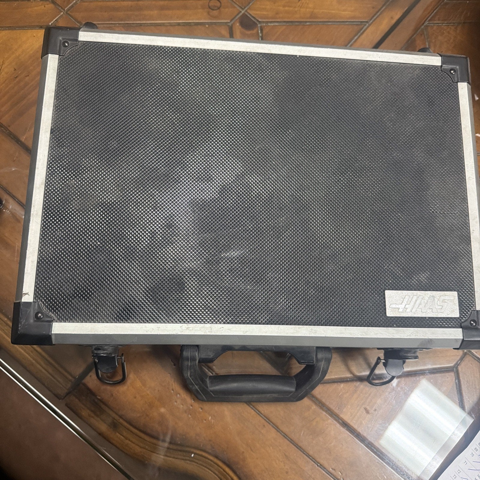 Closed rectangular hard carrying case with textured black surface, silver metal edges, reinforced corners, black plastic handle, and "HAAS" logo plate on a wooden table.