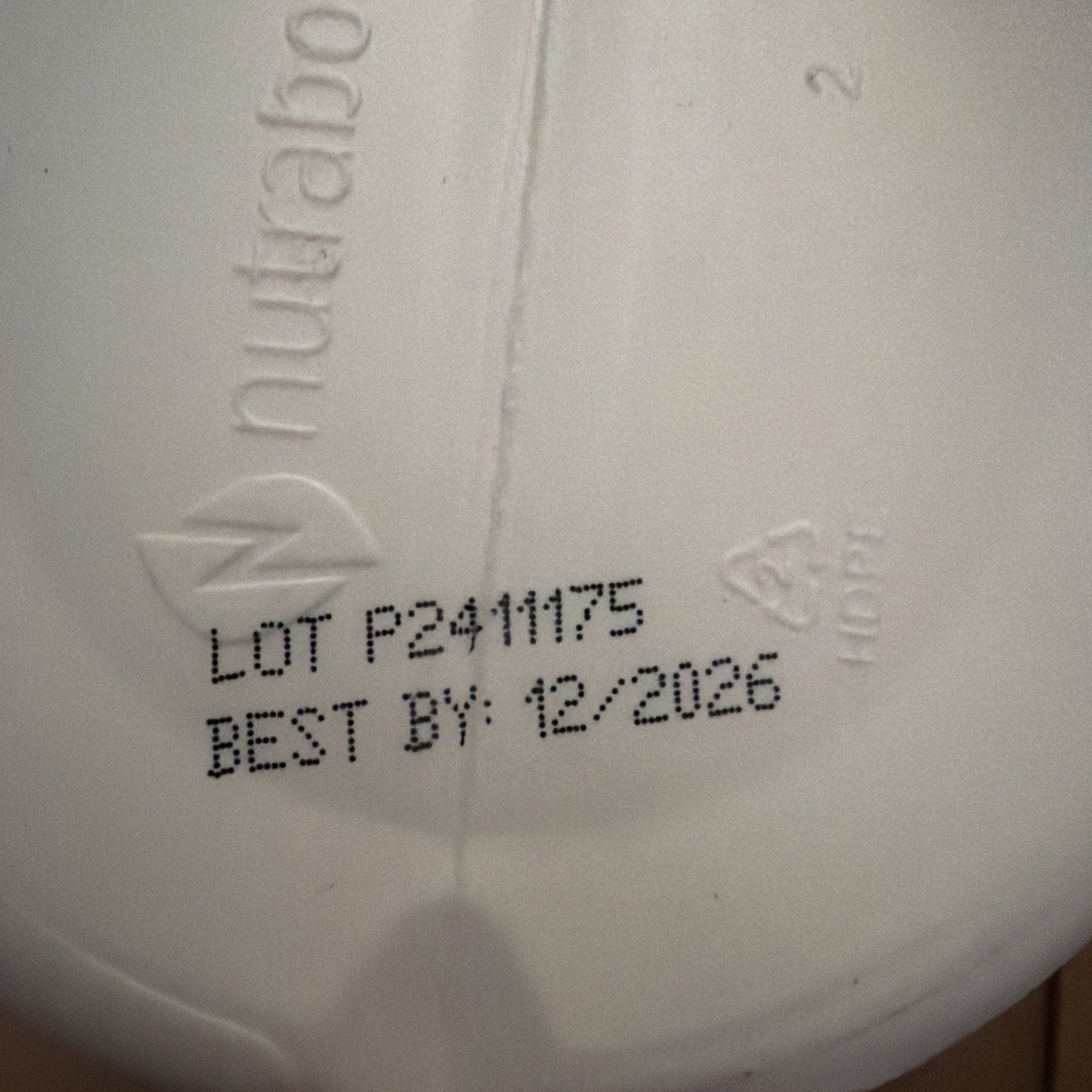 Close-up of a white container bottom with embossed brand name "nutra" and recycling symbol "2 HDPE," showing printed lot number "P2411175" and best-by date "12/2026.