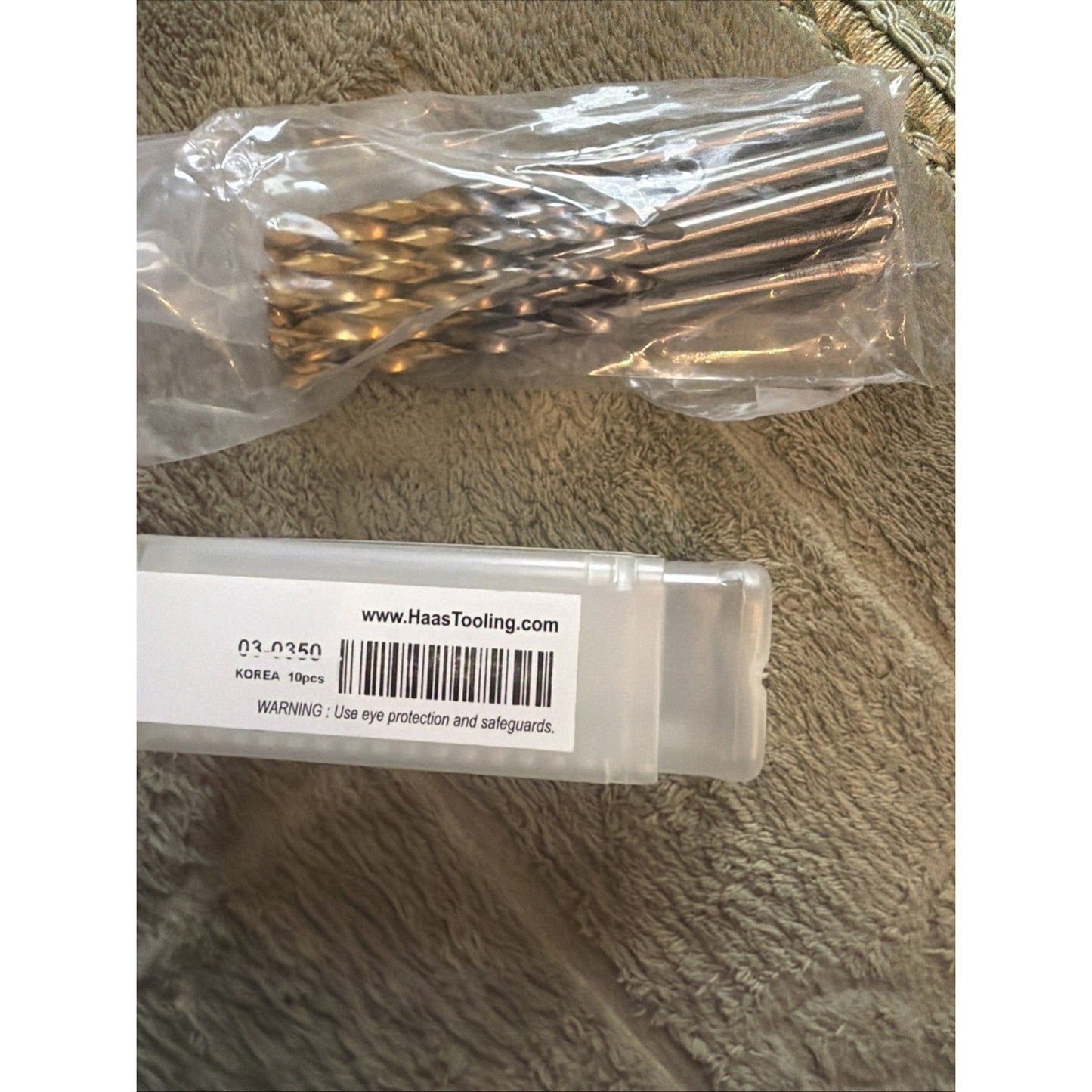 Ten metal twist drill bits with spiral flutes packaged in a clear plastic bag, shown alongside a transparent plastic case labeled with "www.HaasTooling.com" and safety warning, placed on a textured fabric surface.