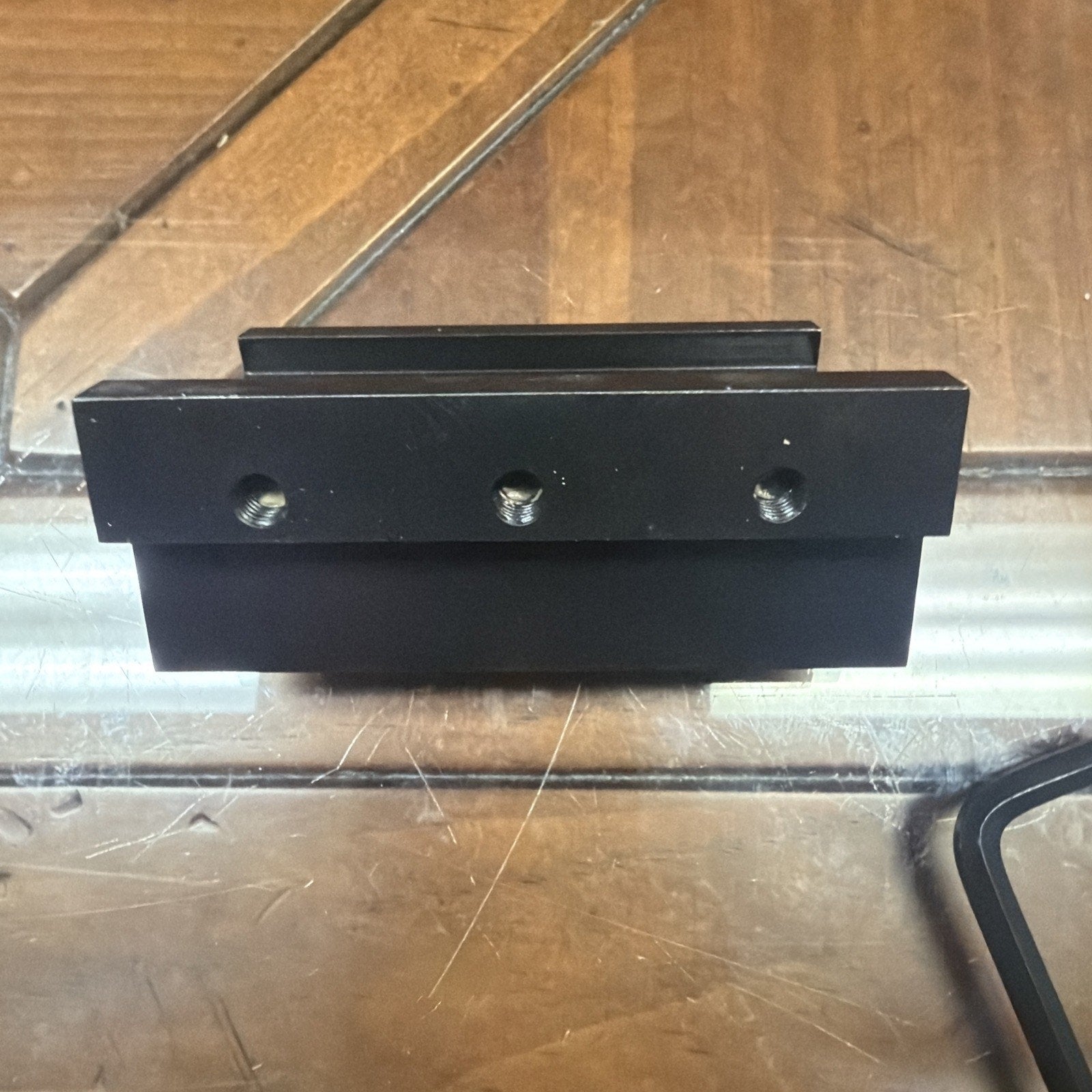 Black rectangular metal clamp with three threaded holes on the front face, placed on a reflective glass surface over a wooden background.