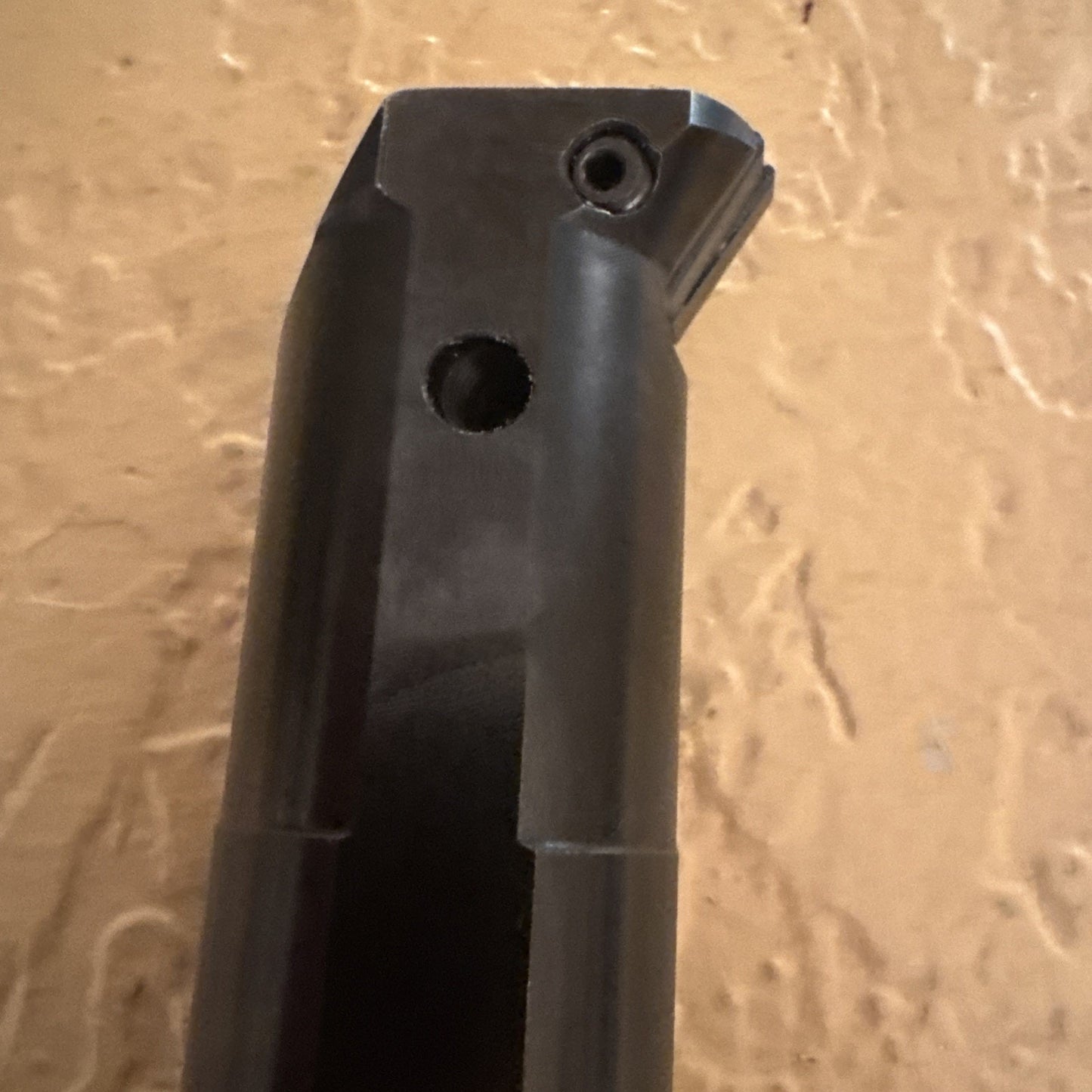 Close-up of a black metal cutting tool tip with a small angled cutting edge secured by a screw, featuring a circular hole below the tip, set against a textured beige background.