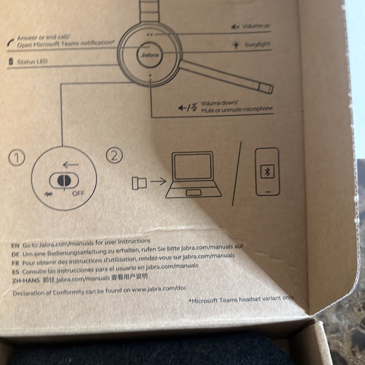 Inside a cardboard box, printed instructions show how to power on a Jabra Bluetooth headset and pair it with a computer or phone using a USB dongle, including volume and call controls.