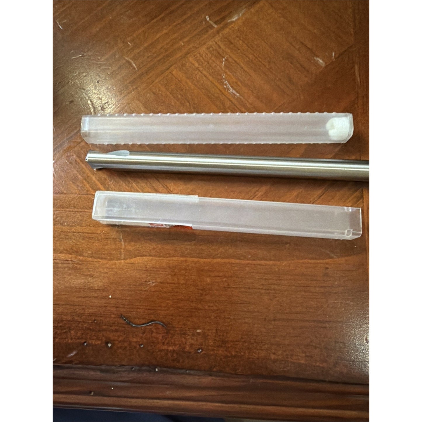Silver metal rod tool with a grooved cutting tip, accompanied by two clear plastic protective cases, resting on a wooden surface with visible grain and minor scratches.