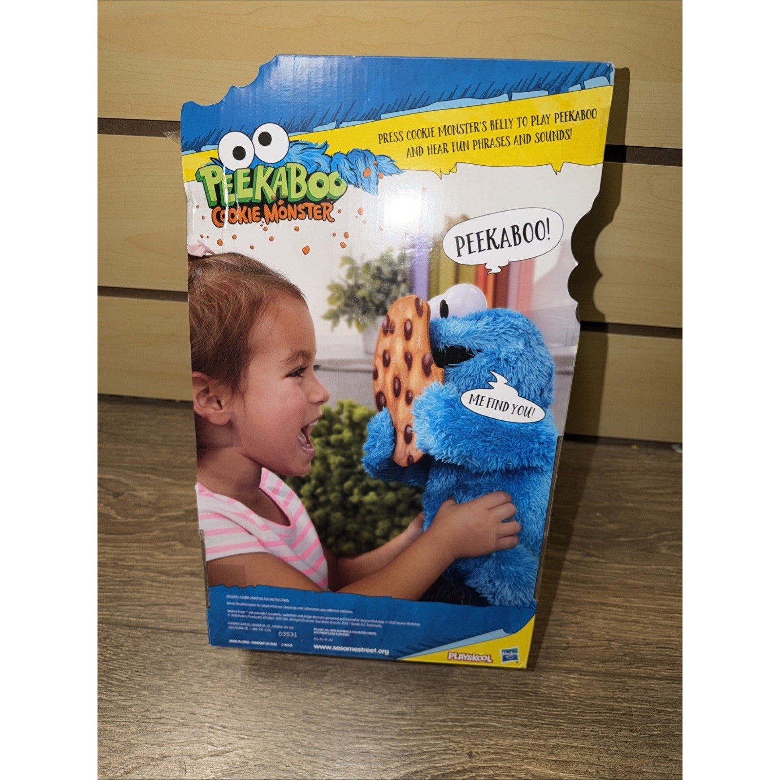 Colorful product packaging featuring a young girl interacting with a blue Cookie Monster plush toy holding a chocolate chip cookie, with playful speech bubbles saying "Peekaboo!" and "Me find you!