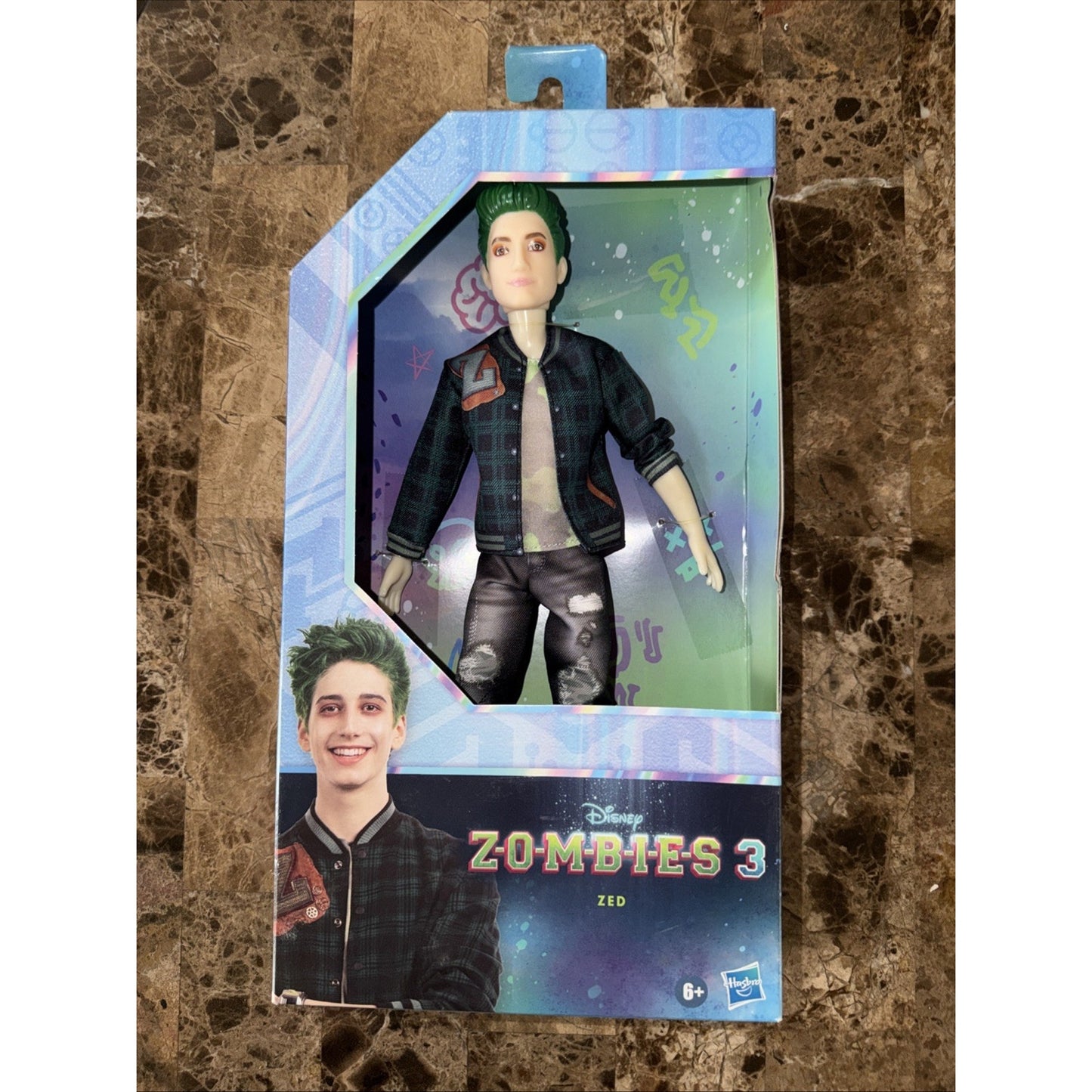 Action figure of Zed from Disney Zombies 3 wearing a black and green plaid jacket and ripped black pants inside a rectangular box with colorful graphics and logo.