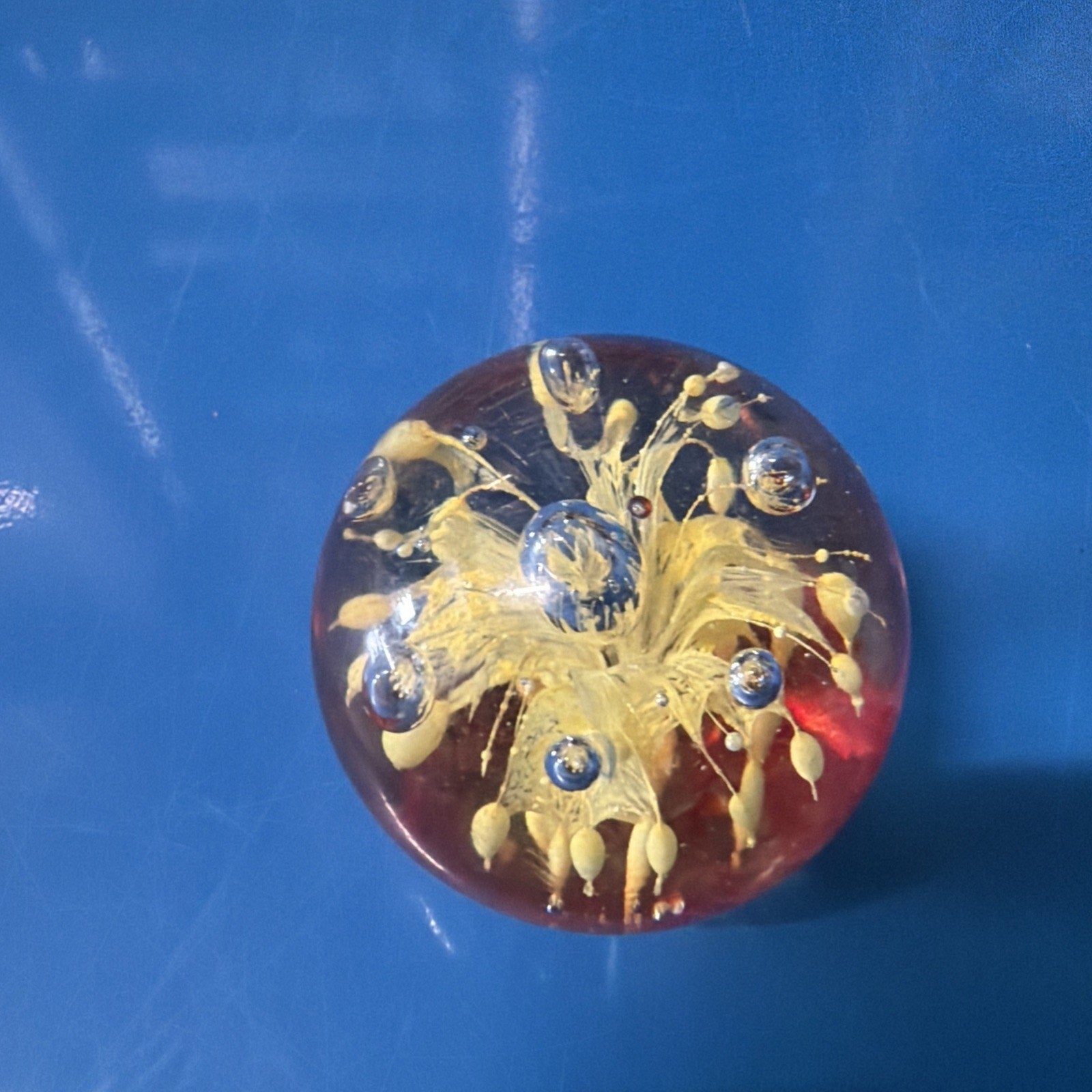 Clear round glass paperweight with a yellow floral design and multiple air bubbles inside, set against a blue surface background.