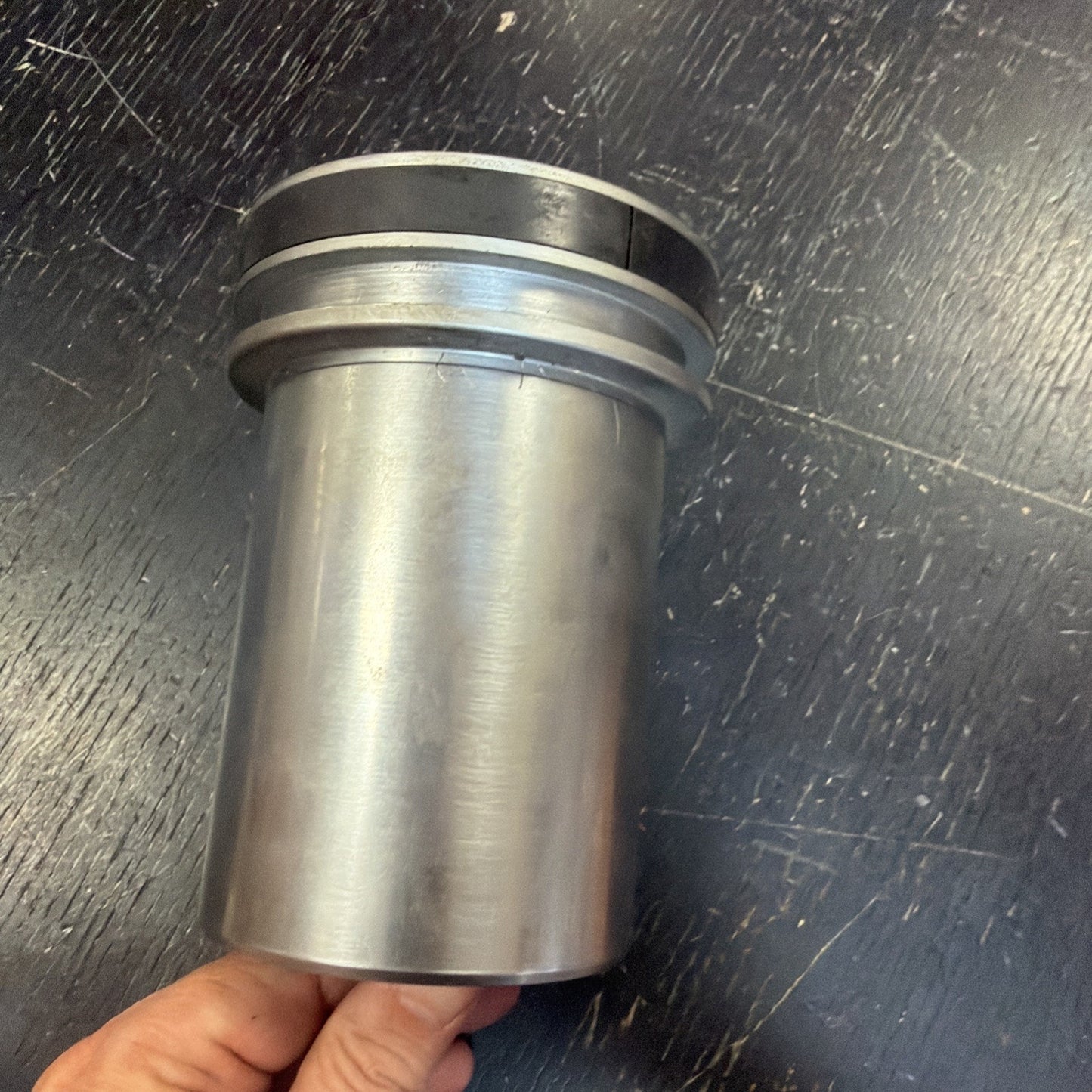 Hand holding a cylindrical metal object with a smooth surface and a grooved rim at the top, positioned against a dark textured background.