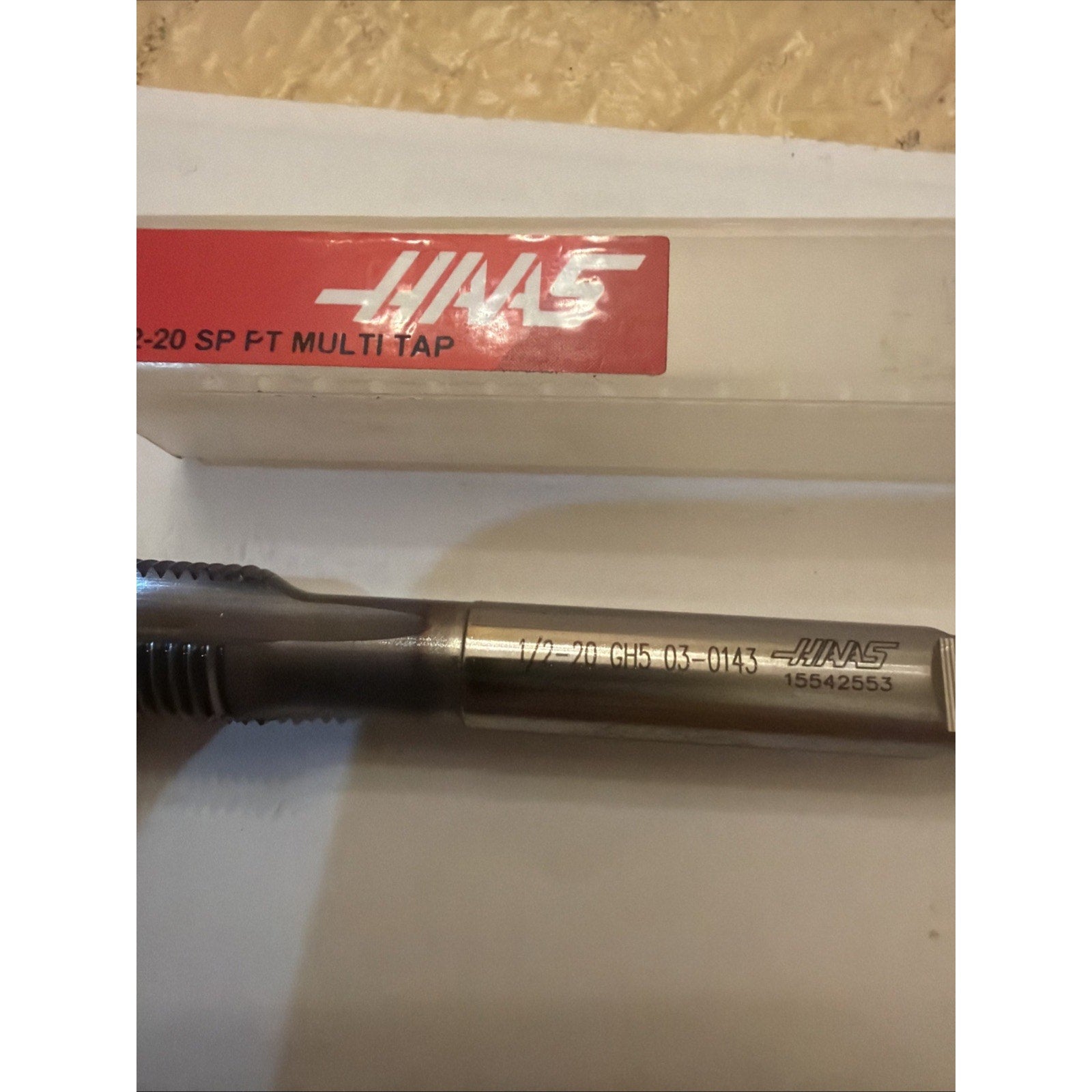 Close-up of a metallic threading tap with engraved size and product numbers along the shank, placed on a white surface next to a clear plastic case with a red Haas label.