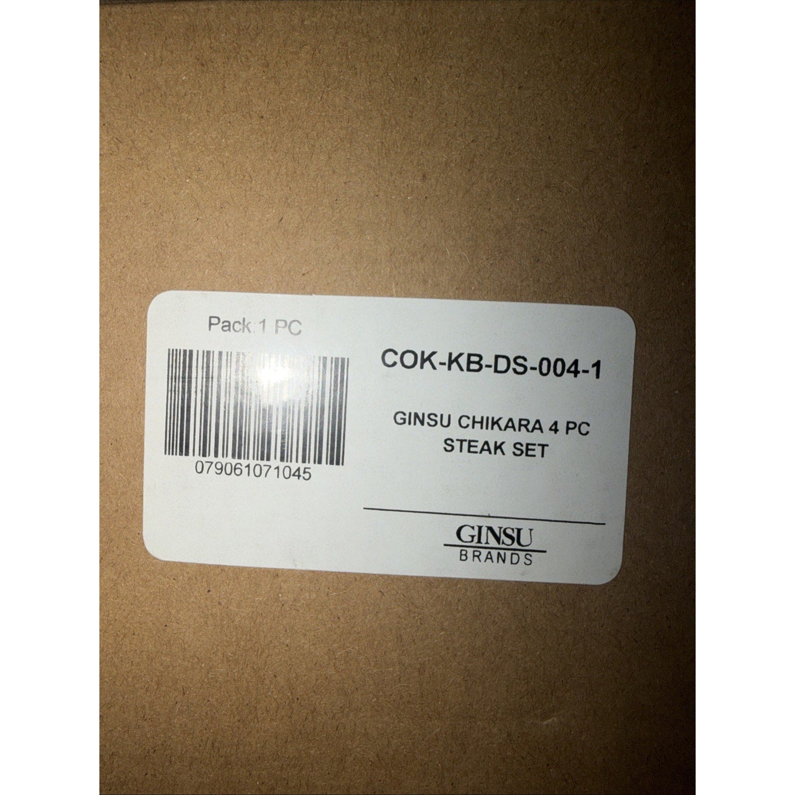 Close-up of a white product label on brown packaging displaying a barcode, product code, and text indicating a four-piece Ginsu Chikara steak knife set from Ginsu Brands.