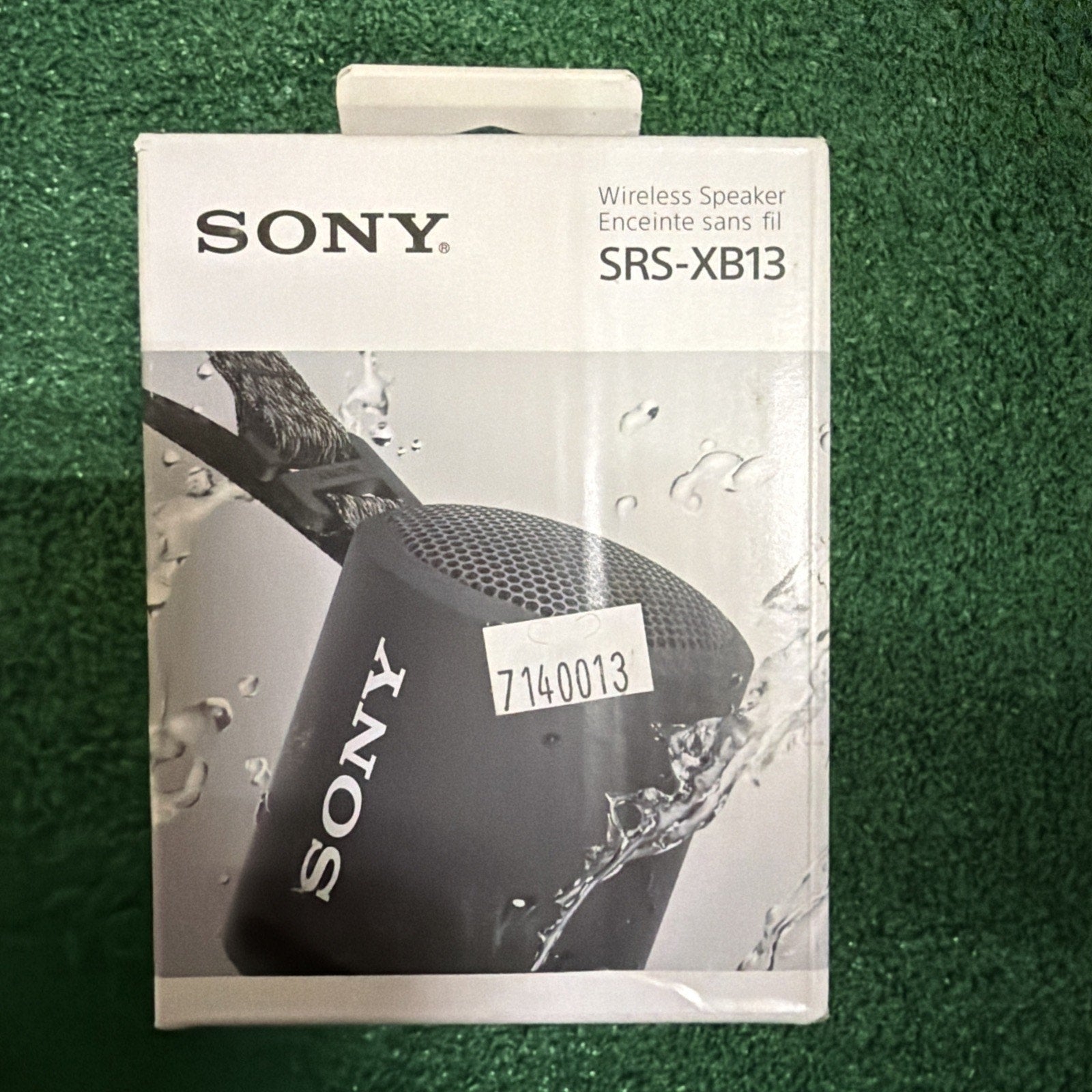 Box packaging of a black Sony SRS-XB13 wireless speaker with water splashing around it, displayed against a green textured background.