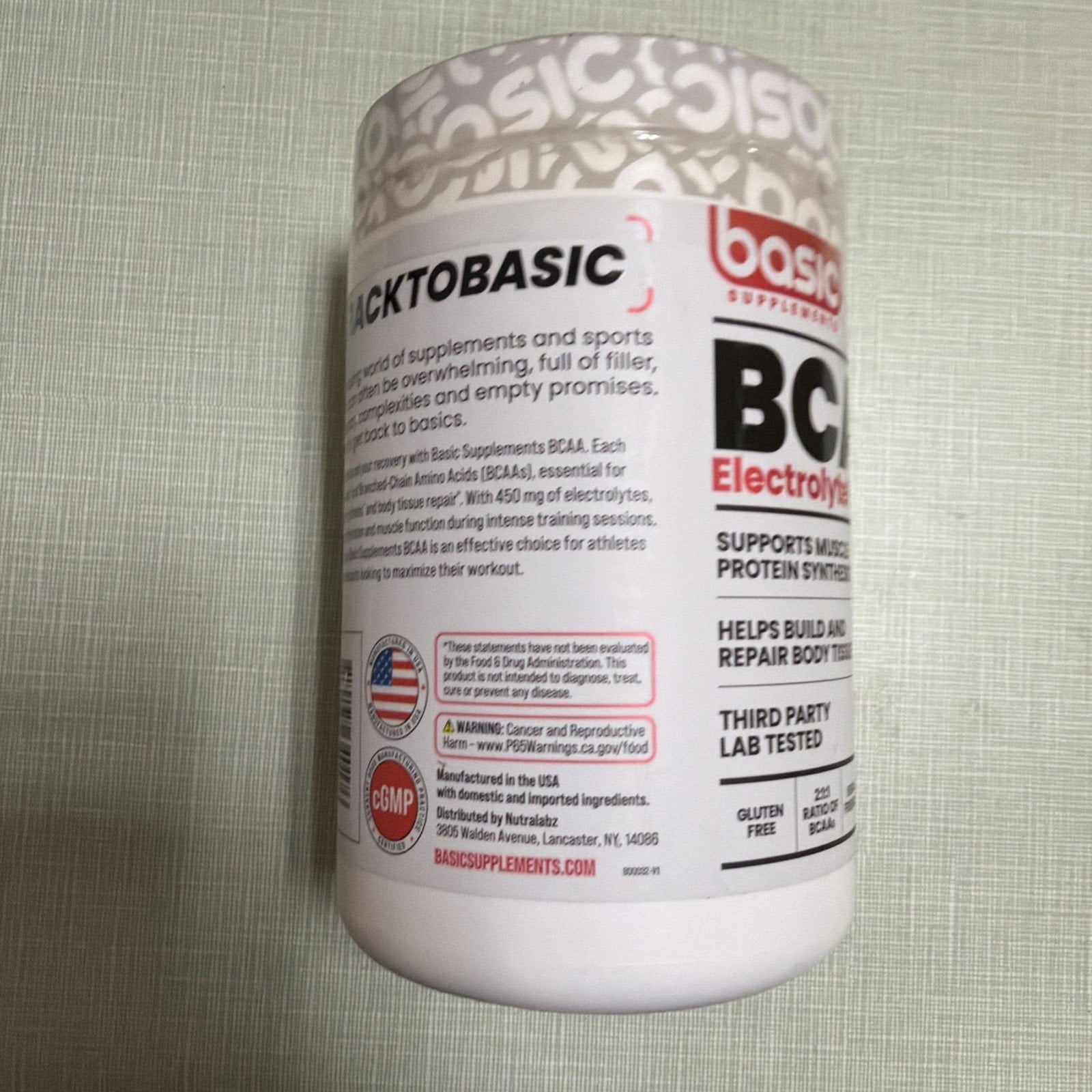 White supplement container with red and black text displaying product benefits for muscle support and protein synthesis, third-party lab testing, gluten-free and batch-tested labels, along with manufacturing and safety information on the side.
