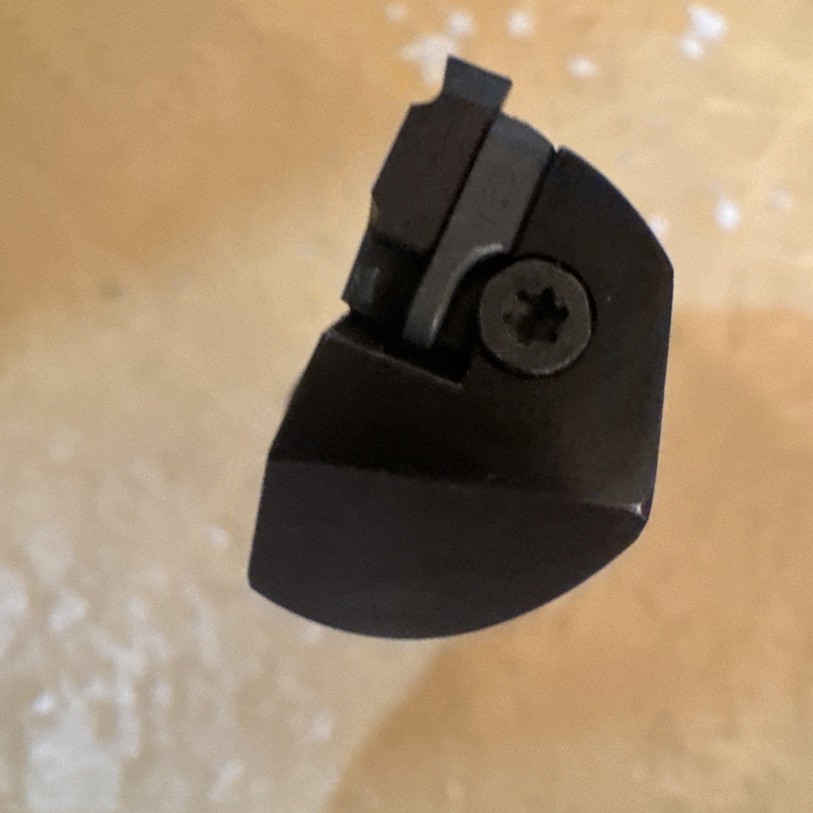 Top-down close-up of a dark metal cutting tool tip with a small rectangular insert secured by a screw, set against a light beige textured background.