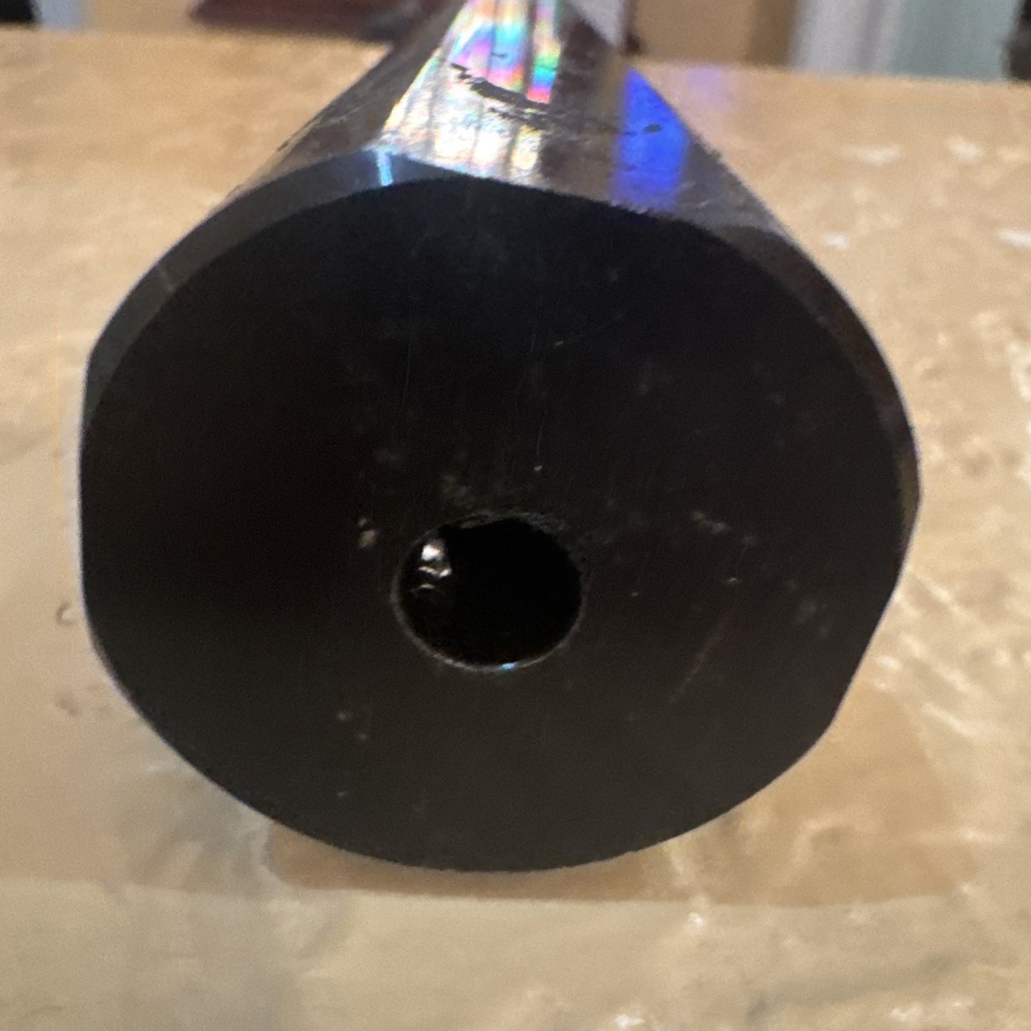 Close-up view of a black cylindrical metal object with a hexagonal outer shape and a central circular hole, resting on a beige surface.