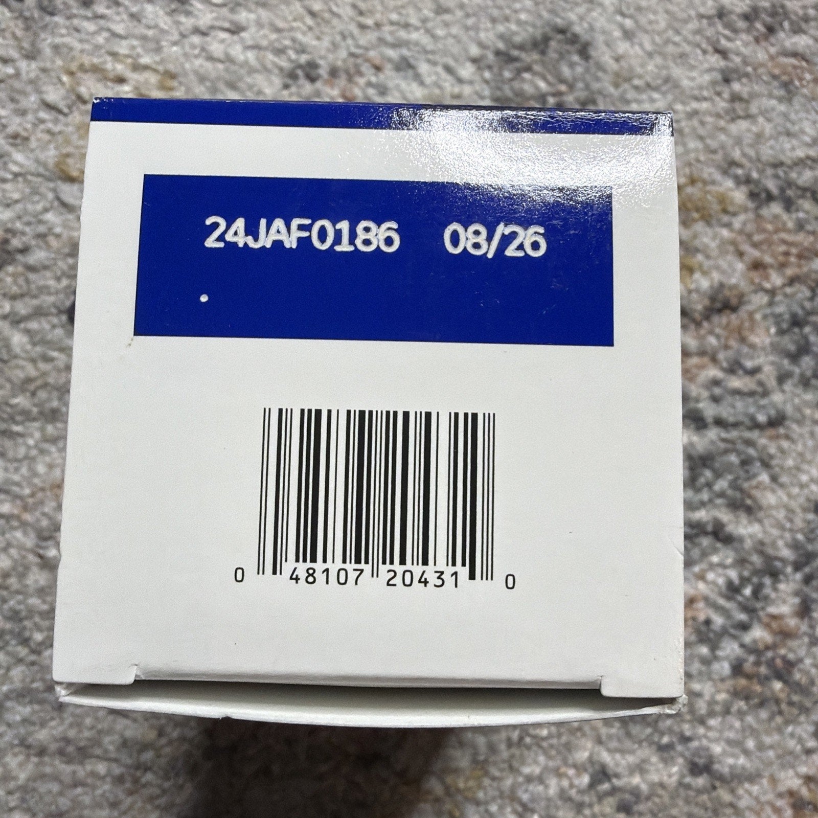 White product box bottom showing a blue label with code 24JAF0186 and expiration date 08/26 above a barcode on a textured gray surface.
