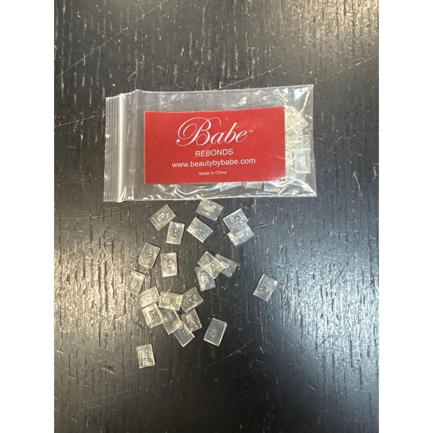 Small clear rectangular lash extension rebonding strips scattered on a dark textured surface, with a resealable plastic bag featuring a red label and white script branding.