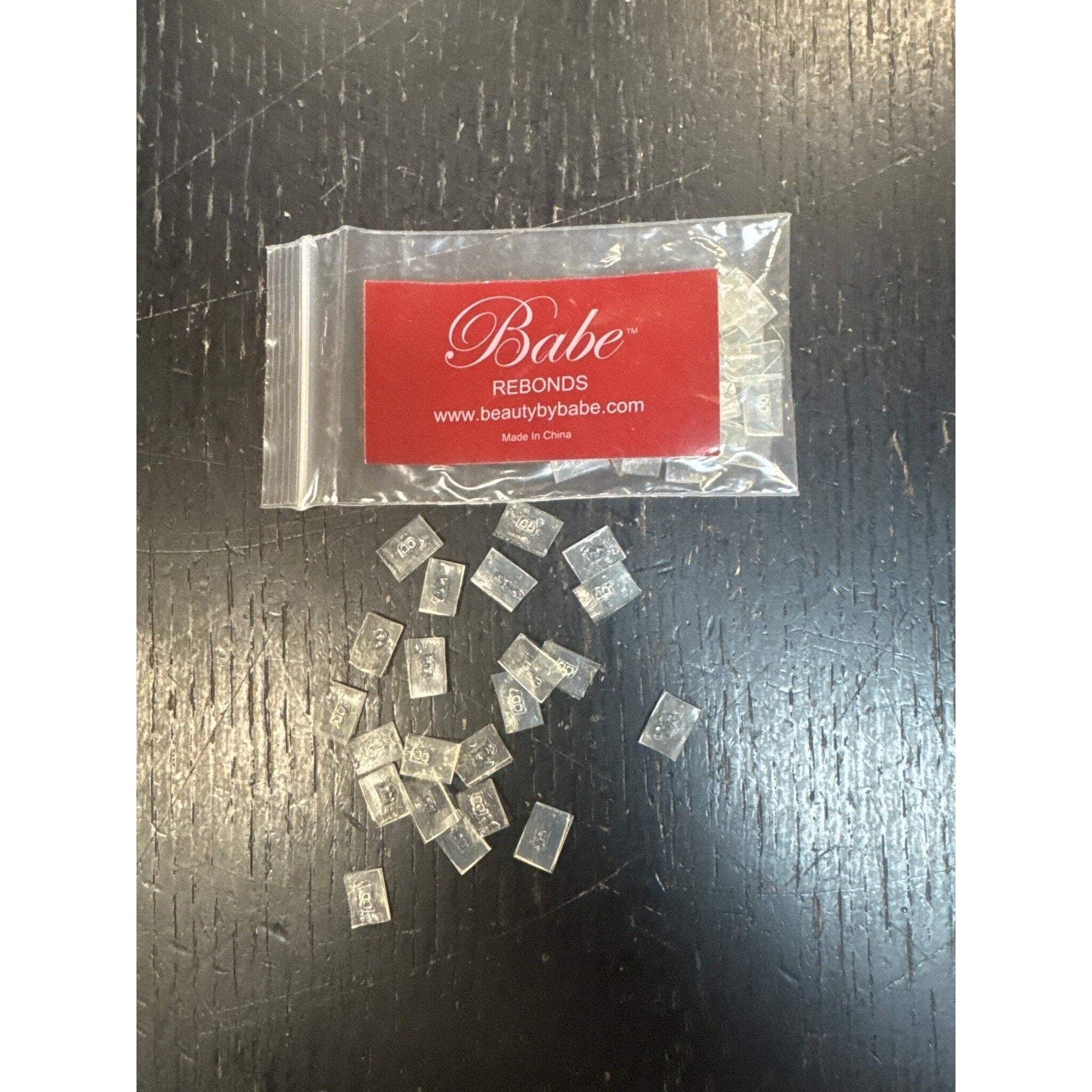 Small clear rectangular lash extension rebonding strips scattered on a dark textured surface, with a resealable plastic bag featuring a red label and white script branding.