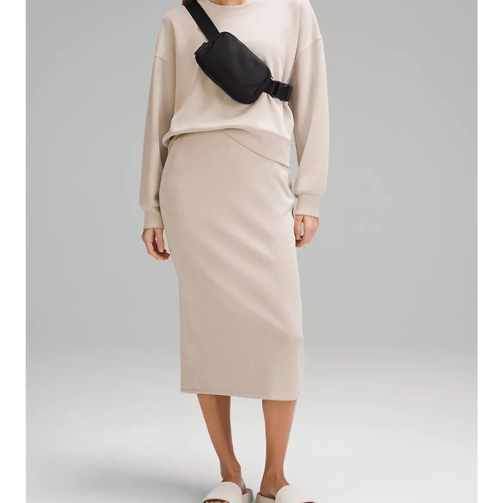 Person wearing a beige long-sleeve top and matching midi skirt paired with white slide sandals, accessorized with a black crossbody bag against a plain gray background.