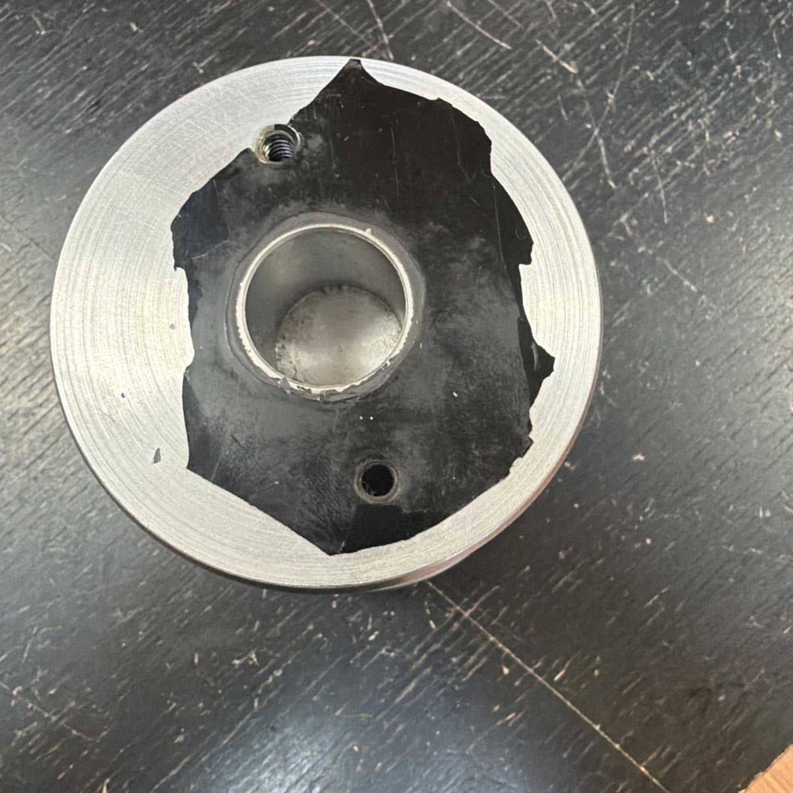 Top-down view of a circular metal component with a central hollow cylinder and two threaded holes, partially covered by a worn black protective tape, placed on a dark textured surface.