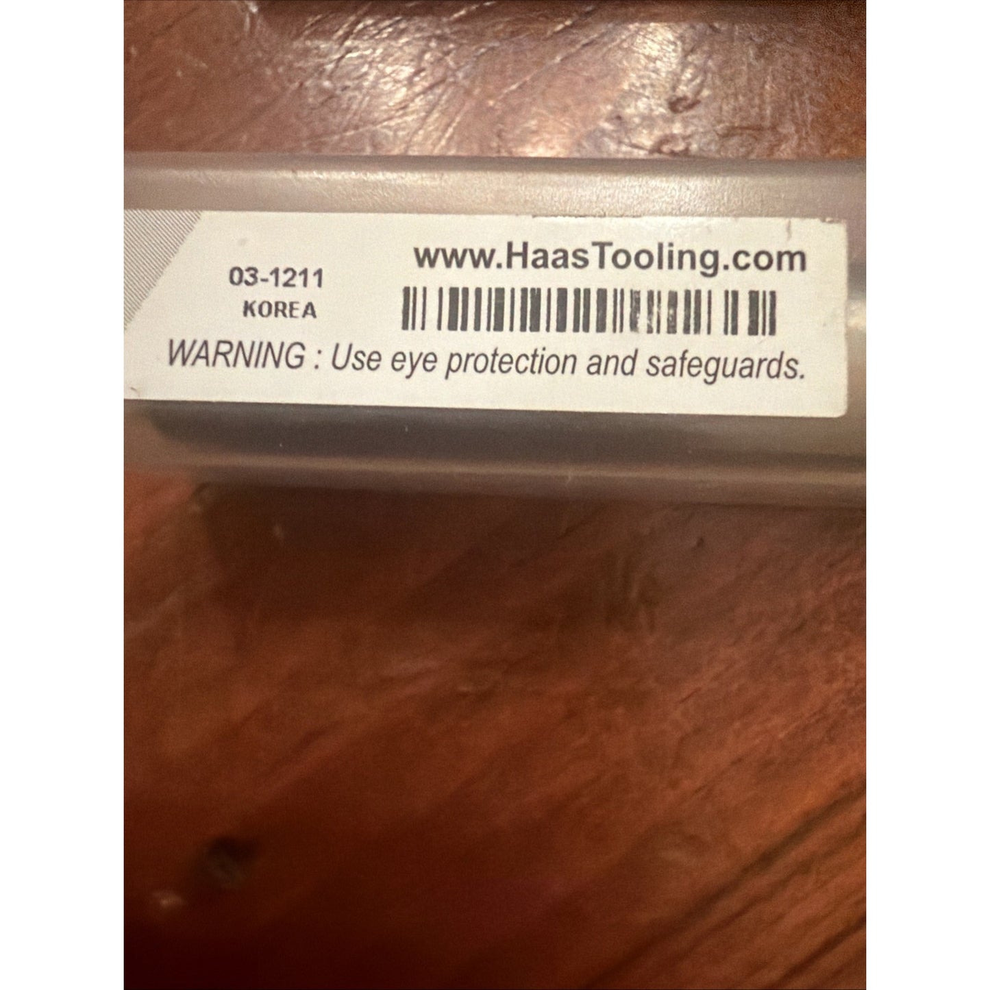 Label on a translucent container showing product code 03-1211, origin Korea, website URL HaasTooling.com, barcode, and a safety warning advising the use of eye protection and safeguards.