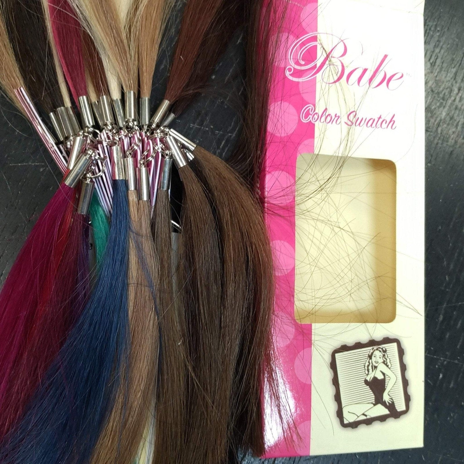 Collection of various colored hair swatches attached to silver metal clips, displayed next to a pink and white packaging card labeled "Babe Color Swatch" on a dark surface.