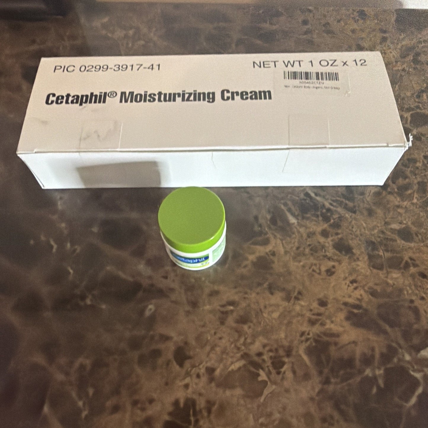 White rectangular box labeled Cetaphil Moisturizing Cream with a small jar featuring a green lid placed in front on a marbled brown surface.