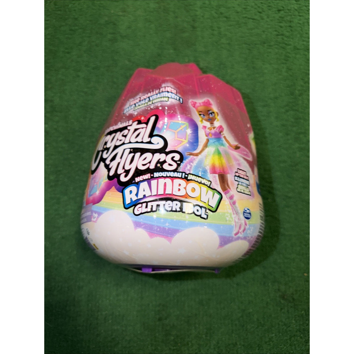 Colorful pink packaging of a Crystal Flyers Rainbow Glitter Doll toy, featuring an illustrated fairy character in a rainbow dress with unicorn ears on a green background.