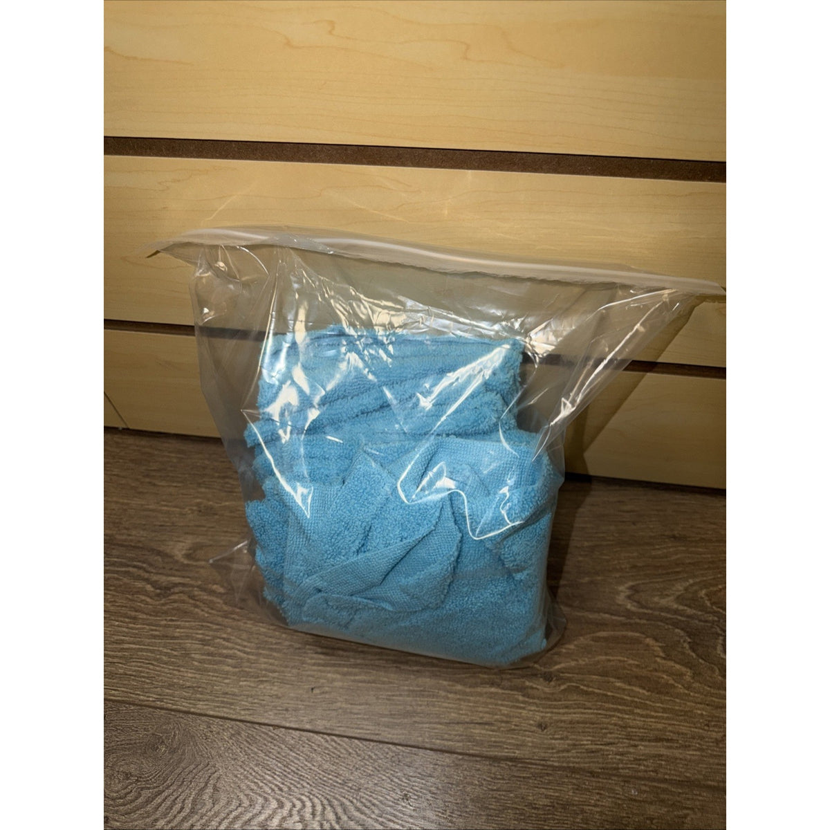 Clear plastic resealable bag containing a folded blue microfiber cloth resting on a wood-textured floor with a light wood panel background.