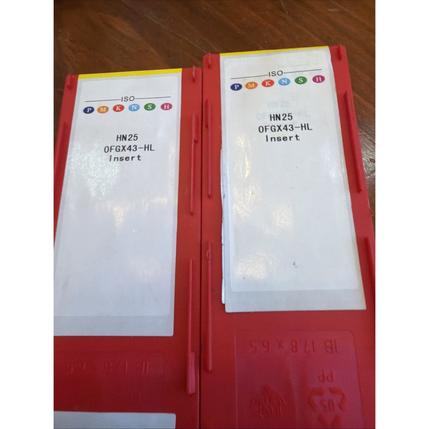 Two red plastic holders with white labels showing colorful ISO classification symbols and text reading "HN25 0FGX43-HL Insert" placed on a wooden surface.