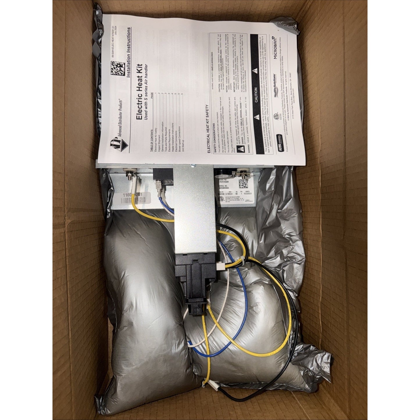 Open brown cardboard box containing an electric heat kit with coiled insulated silver tubing and attached wiring, accompanied by a printed instruction manual.