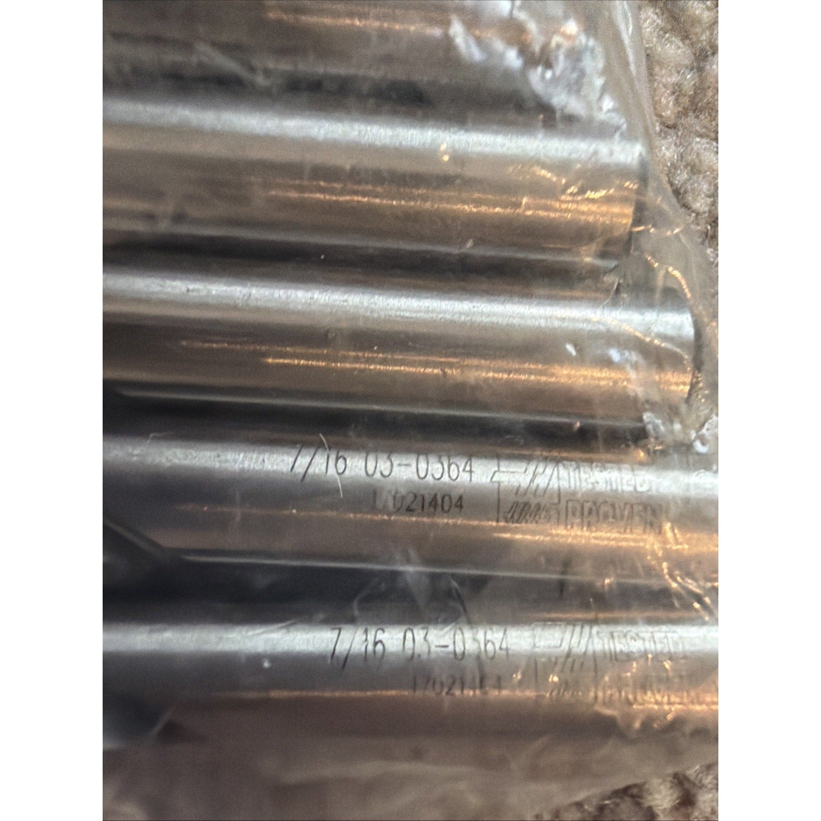 Close-up of several metal cylindrical rods with engraved alphanumeric codes and batch numbers, partially wrapped in clear plastic on a textured surface.