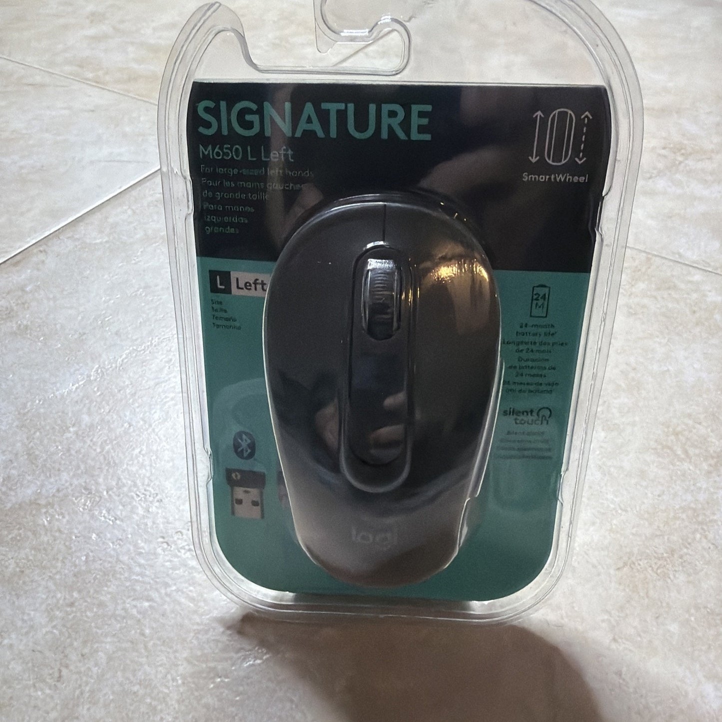 Black wireless computer mouse in clear plastic packaging labeled "Signature M650 L Left" with features including a smart wheel, Bluetooth connectivity, and silent touch technology.