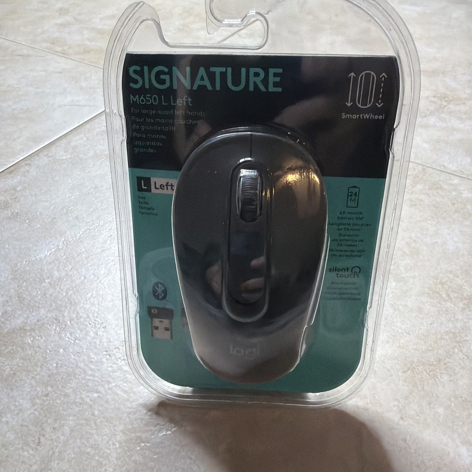 Black wireless computer mouse in clear plastic packaging labeled "Signature M650 L Left" with features including a smart wheel, Bluetooth connectivity, and silent touch technology.