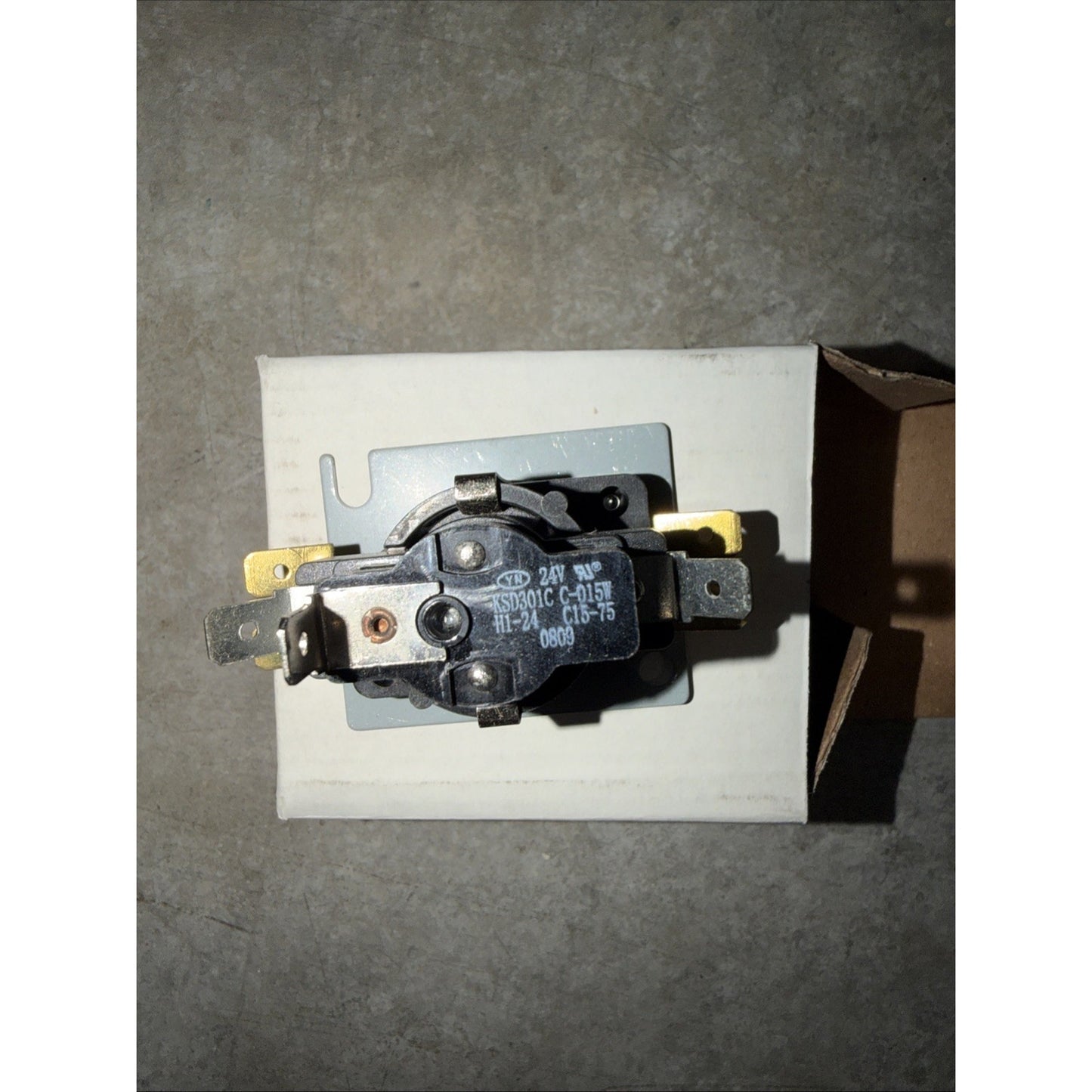 Black electrical component with metal terminals mounted on a small metal plate, placed on a white cardboard box against a gray concrete surface.