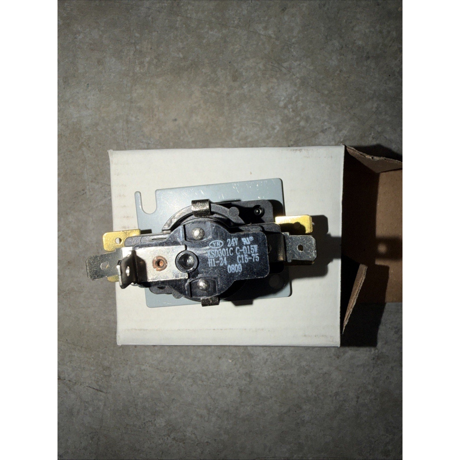 Black electrical component with metal terminals mounted on a small metal plate, placed on a white cardboard box against a gray concrete surface.
