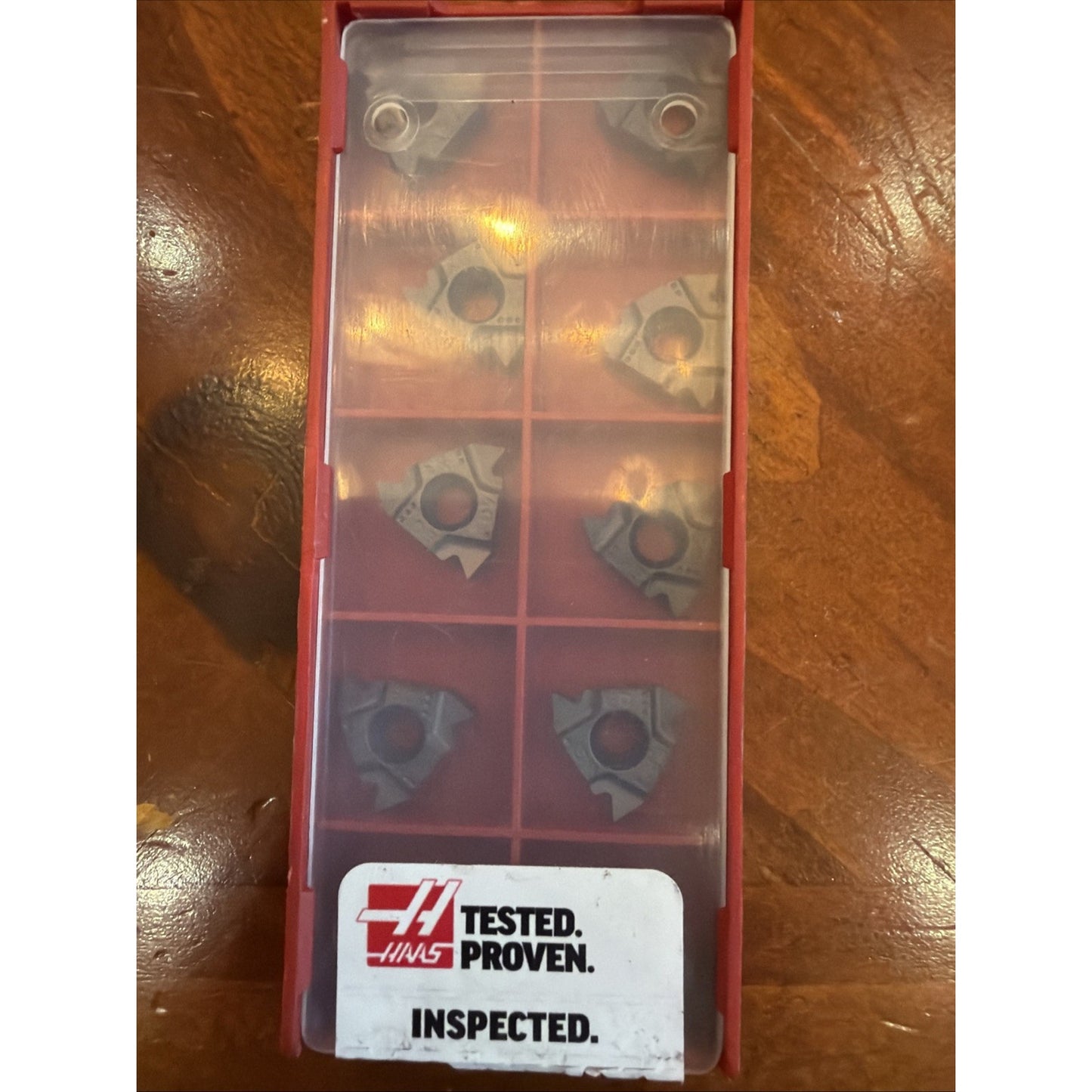 Red plastic storage case with a translucent cover containing eight uniquely shaped metal inserts arranged in two columns, labeled with a Haas logo and text reading "TESTED. PROVEN. INSPECTED." on a wooden surface.