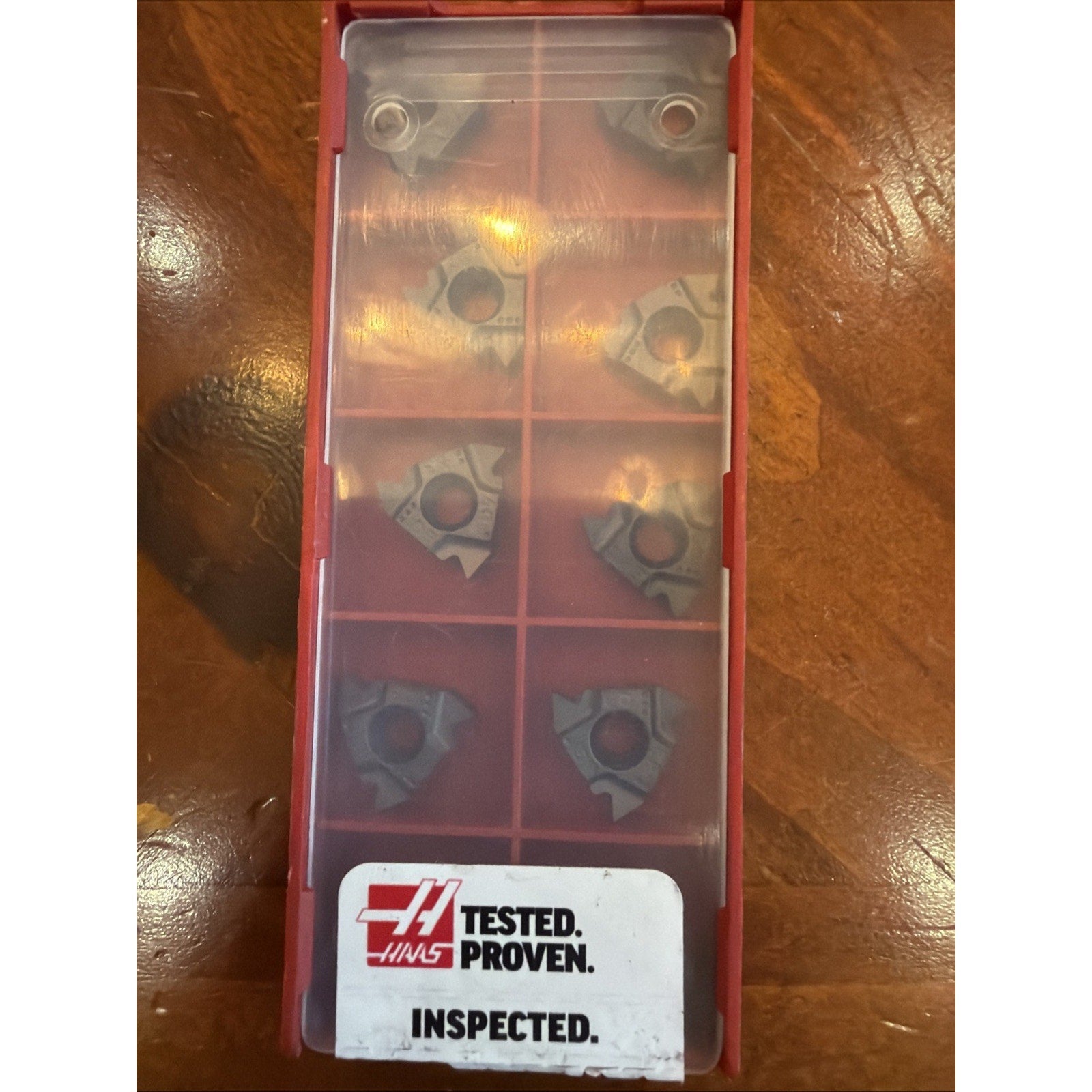 Red plastic storage case with a translucent cover containing eight uniquely shaped metal inserts arranged in two columns, labeled with a Haas logo and text reading "TESTED. PROVEN. INSPECTED." on a wooden surface.