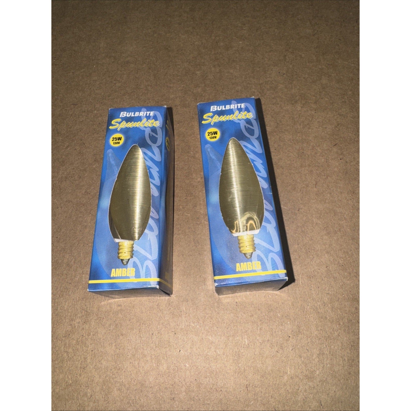 Two Bulbrite Spunlite 25W 130V amber candelabra lightbulbs in blue and yellow retail boxes on brown surface