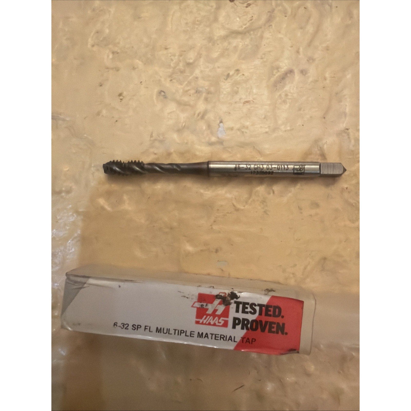 Metal threading tap with spiral flutes and a square drive end, resting on a textured surface above a white and red labeled plastic storage case marked with testing and product details.
