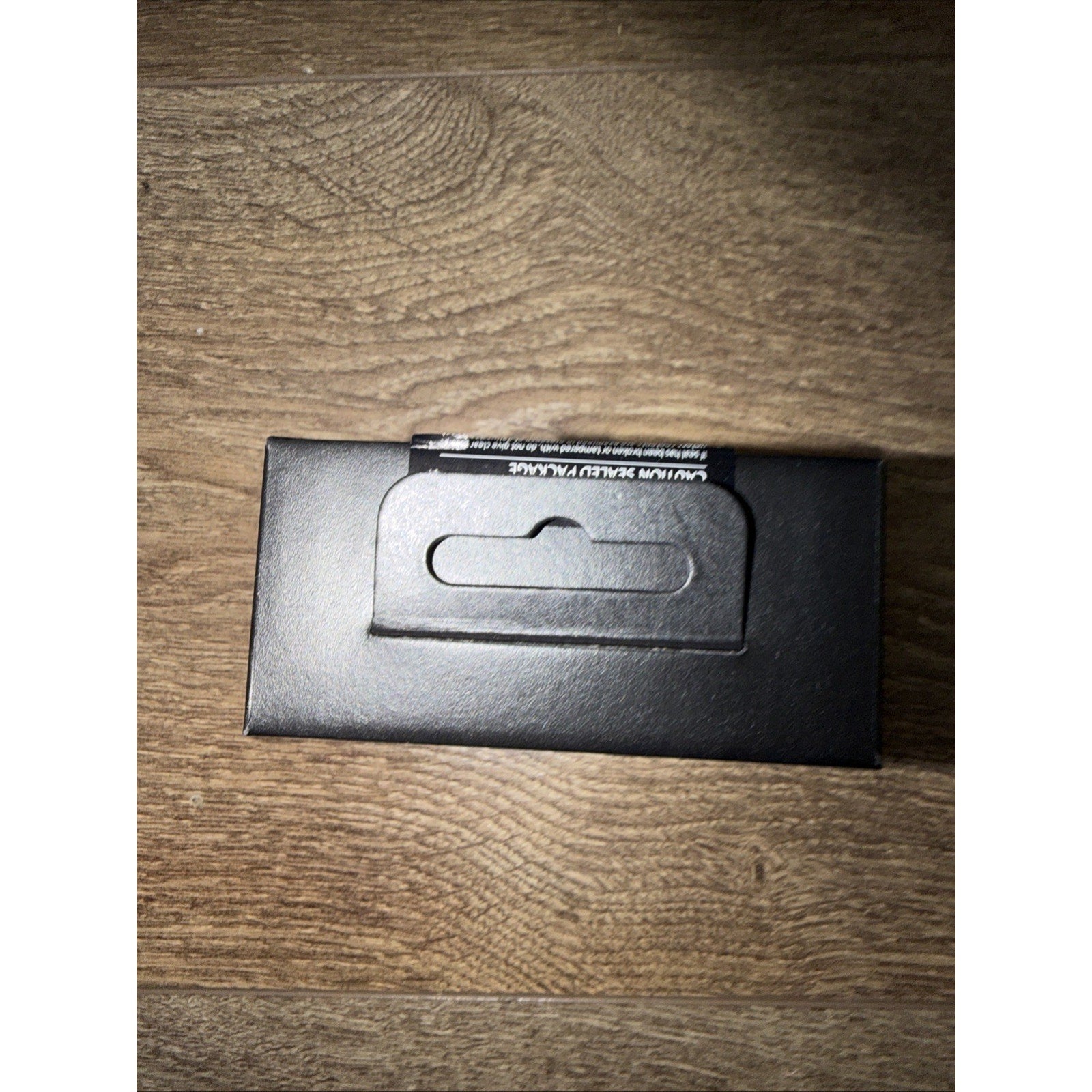 Black rectangular cardboard box with a foldable hanging tab on top, placed on a wooden surface with a textured grain pattern.
