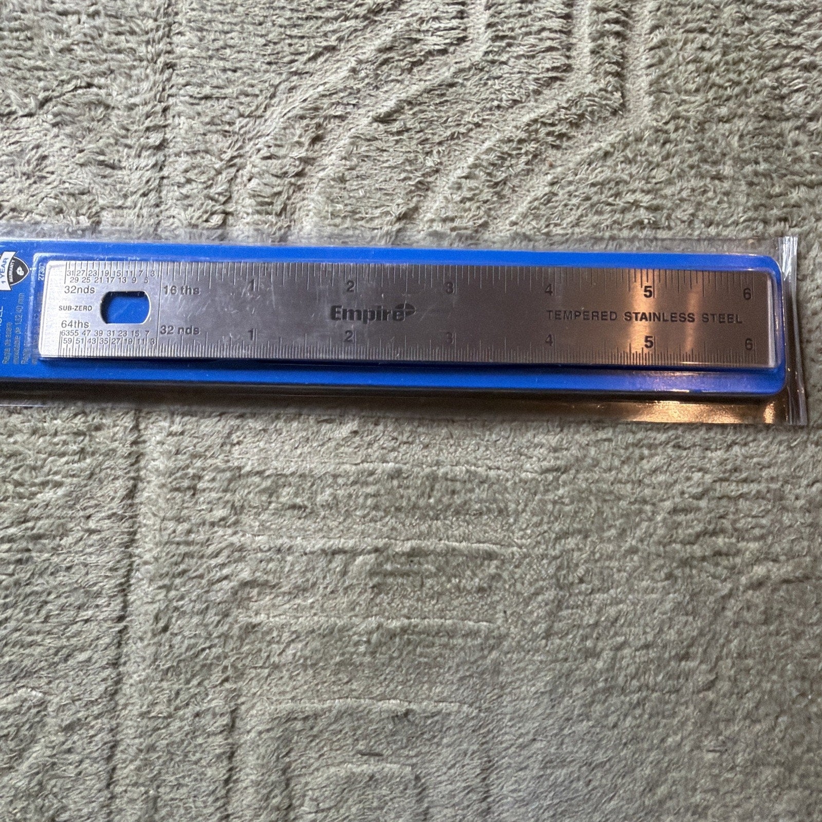 Tempered stainless steel ruler measuring six inches with inch and fractional markings, packaged in a clear plastic sleeve with a blue border, resting on a textured light gray surface.